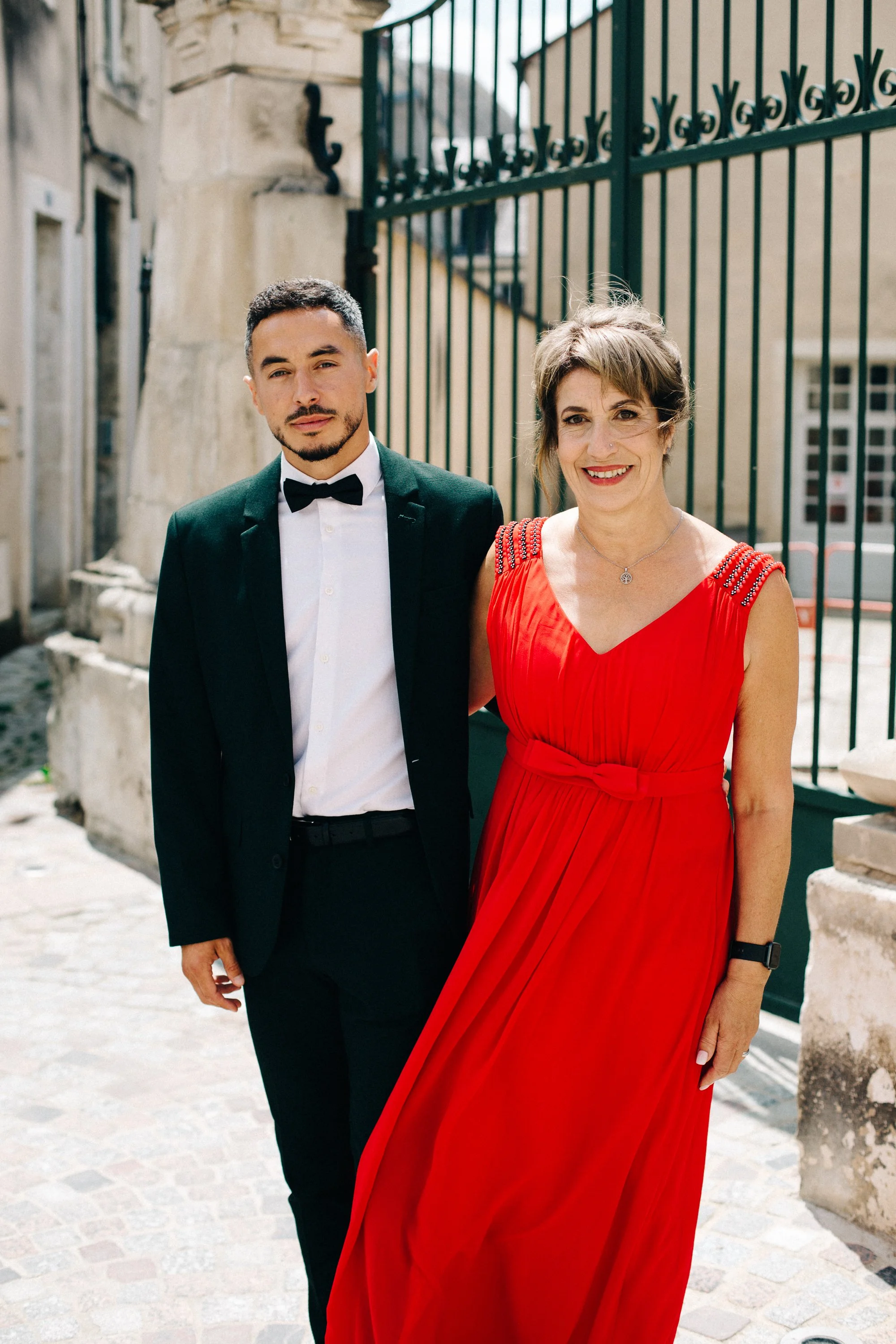 Un homme en costume noir avec une chemise blanche et un nœud papillon, et une femme en robe rouge, posant ensemble devant une grille en fer forgé dans une rue pavée en plein jour.