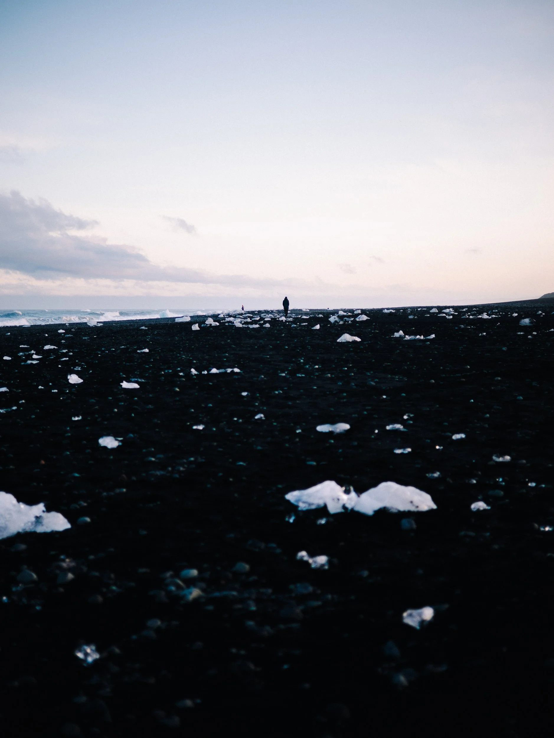 Personne marchant sur une plage noire avec des morceaux de glace, mer agitée en arrière-plan et ciel nuageux, ambiance froide et isolée.