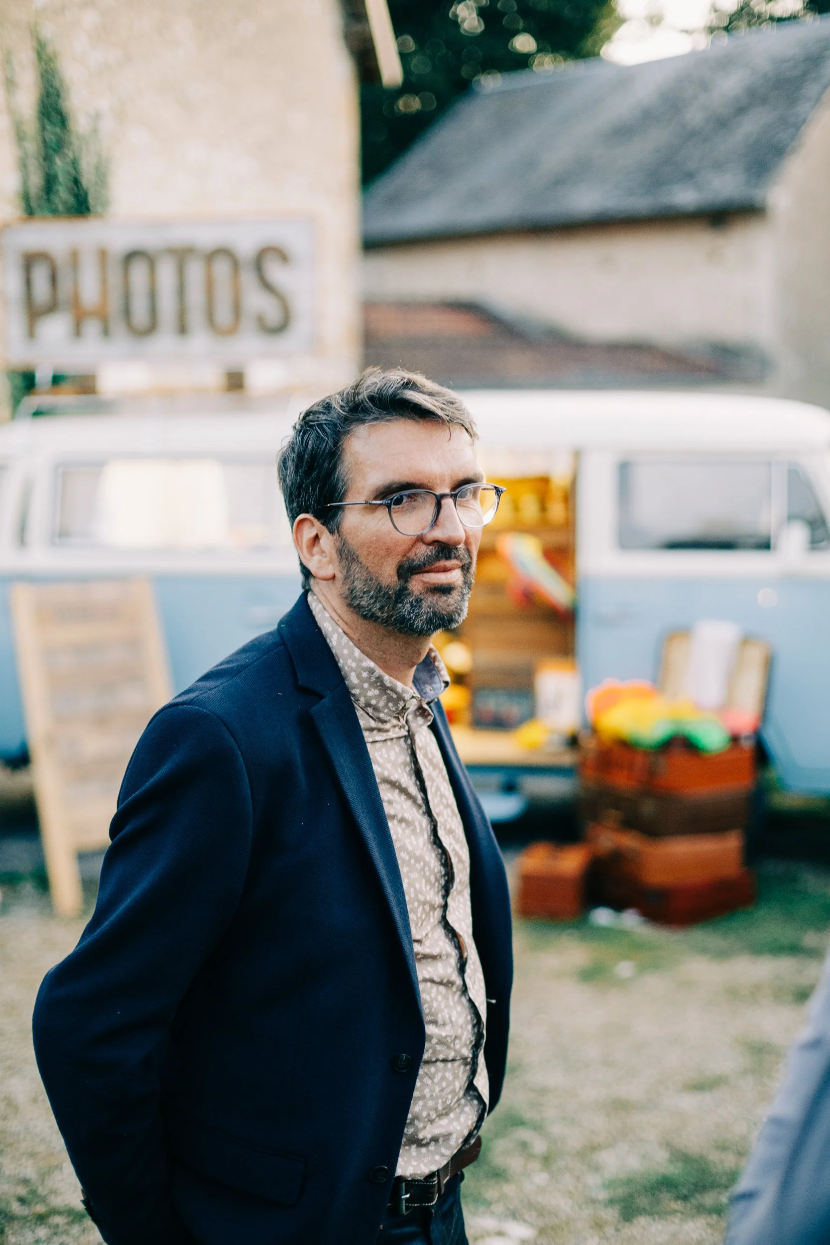 Un homme portant des lunettes, en veste sombre et chemise à motif, se tient devant un camion vintage blanc et blue, avec un panneau 'PHOTOS' en arrière-plan, lors d'une journée en plein air.