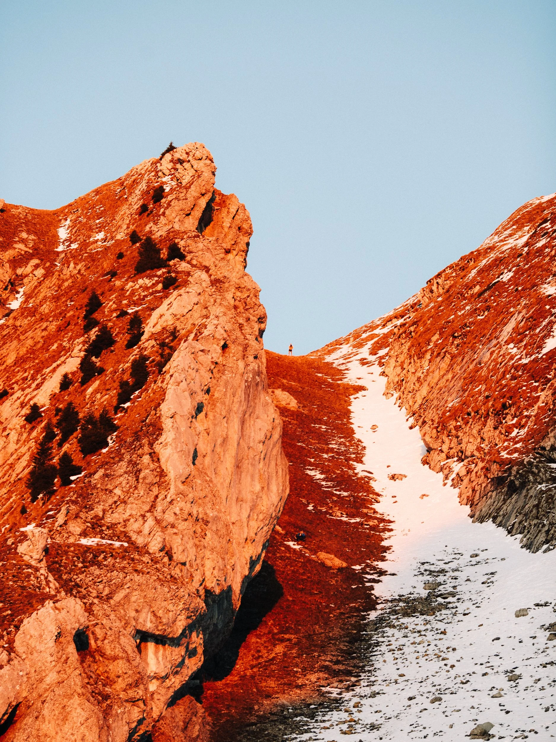 Une vue de montagnes rocheuses avec de la neige au sol et une personne de petite taille au fond.