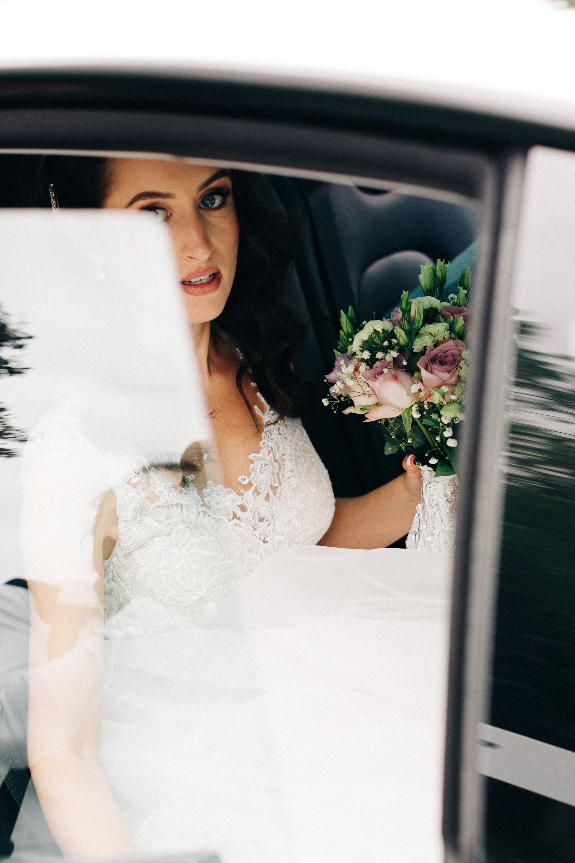 Une femme en robe de mariée tient un bouquet de fleurs et regarde par une fenêtre, vue partielle de son visage.