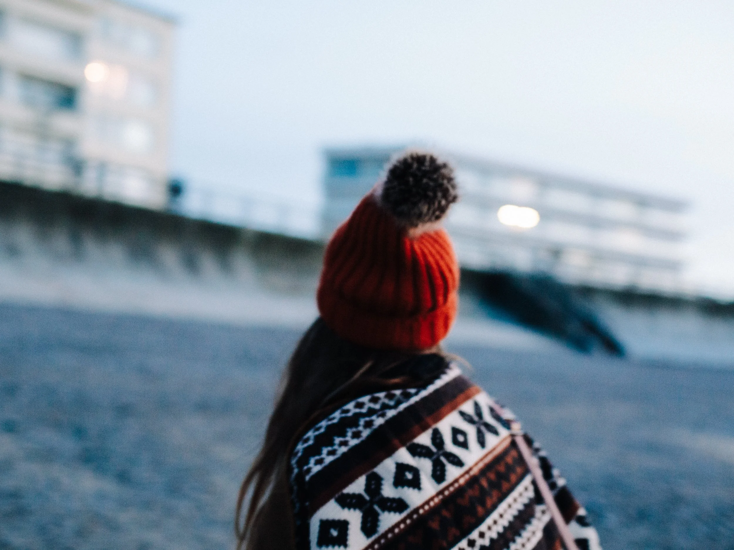 Une personne portant un bonnet en laine à pompon et un pull à motifs géométriques, vue de dos, dans un environnement urbain lors d'une journée froide.