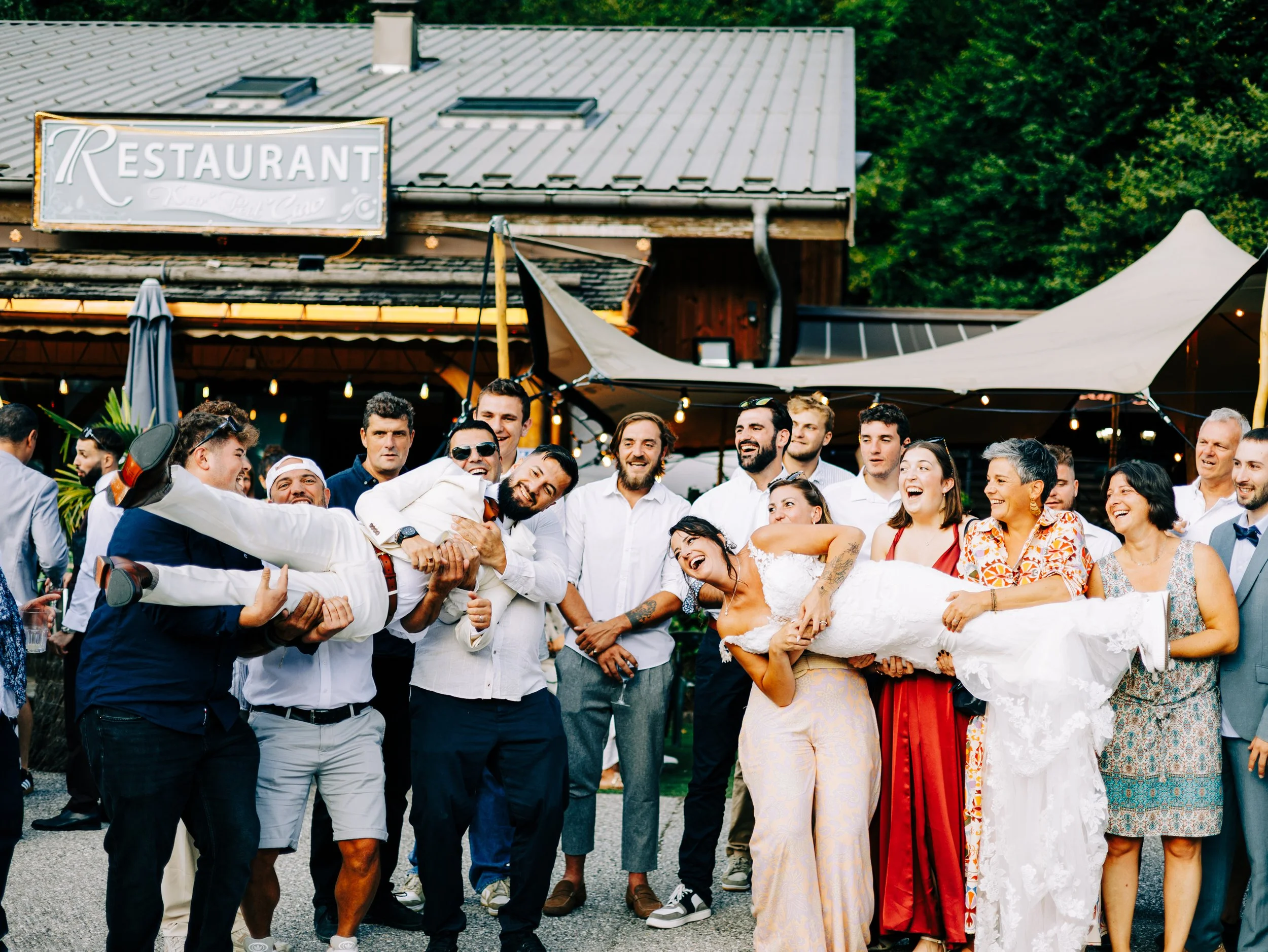 Groupe de personnes souriantes et amicales posant pour une photo en extérieur devant un bâtiment avec un signe indiquant un restaurant. Les gens sont vêtus de façon décontractée, certains en tenues festives, et l'ambiance est joyeuse.
