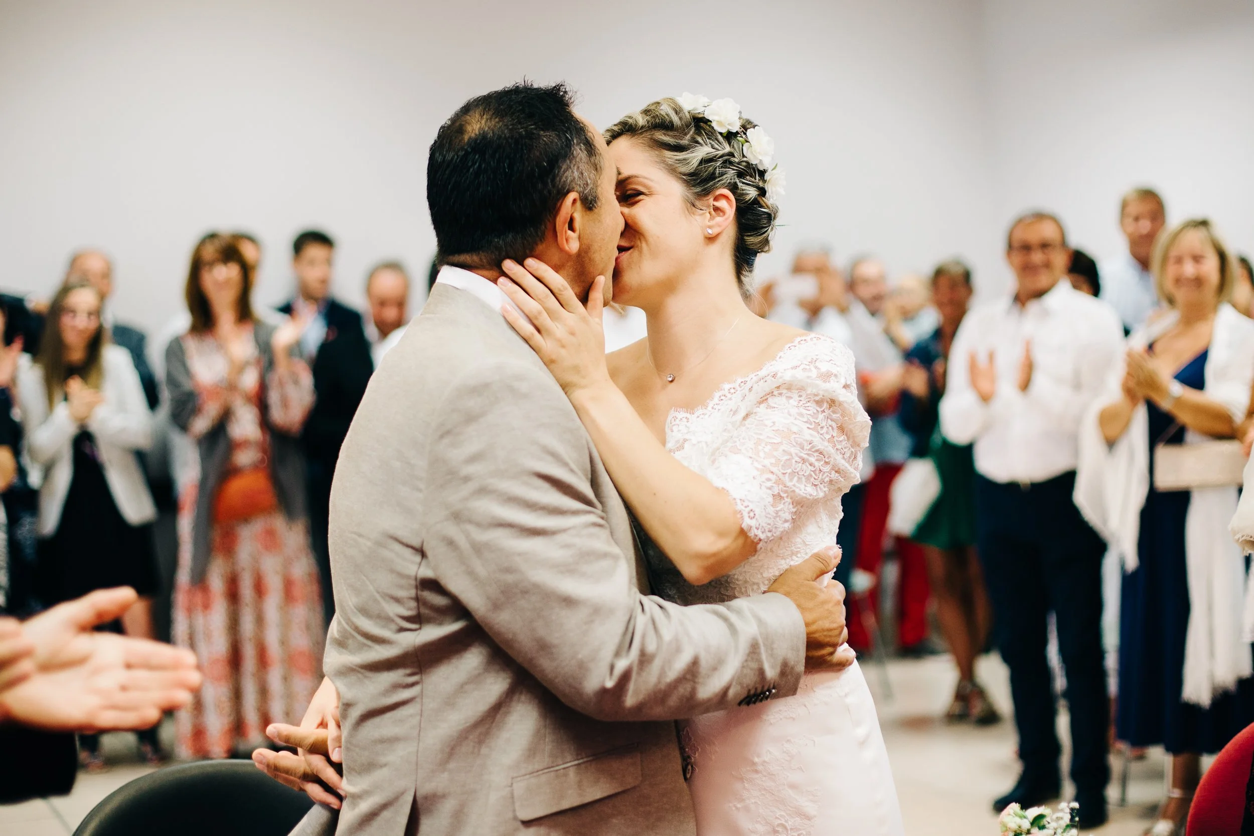 Un couple de mariés s'embrassent lors de leur mariage devant un groupe d'invités. La femme porte une robe de mariée avec des fleurs dans les cheveux, et l'homme porte un costume clair. Les invités applaudissent et sourient en arrière-plan.