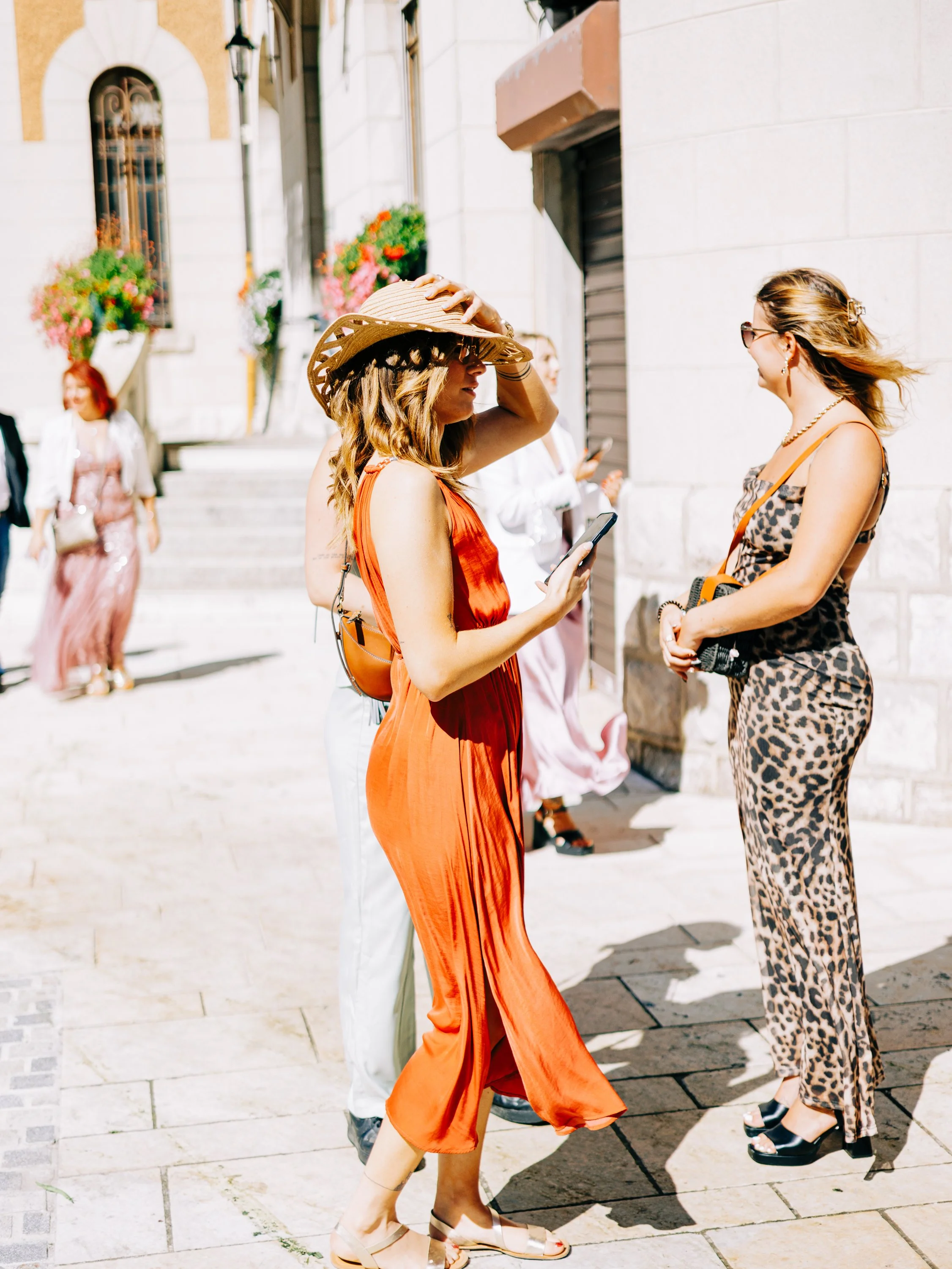 Deux femmes discutent en plein air, l'une porte une robe orange et un grand chapeau, l'autre une robe animal print, toutes deux avec des lunettes de soleil. En arrière-plan, une femme passe devant un bâtiment avec des fleurs en décoration.