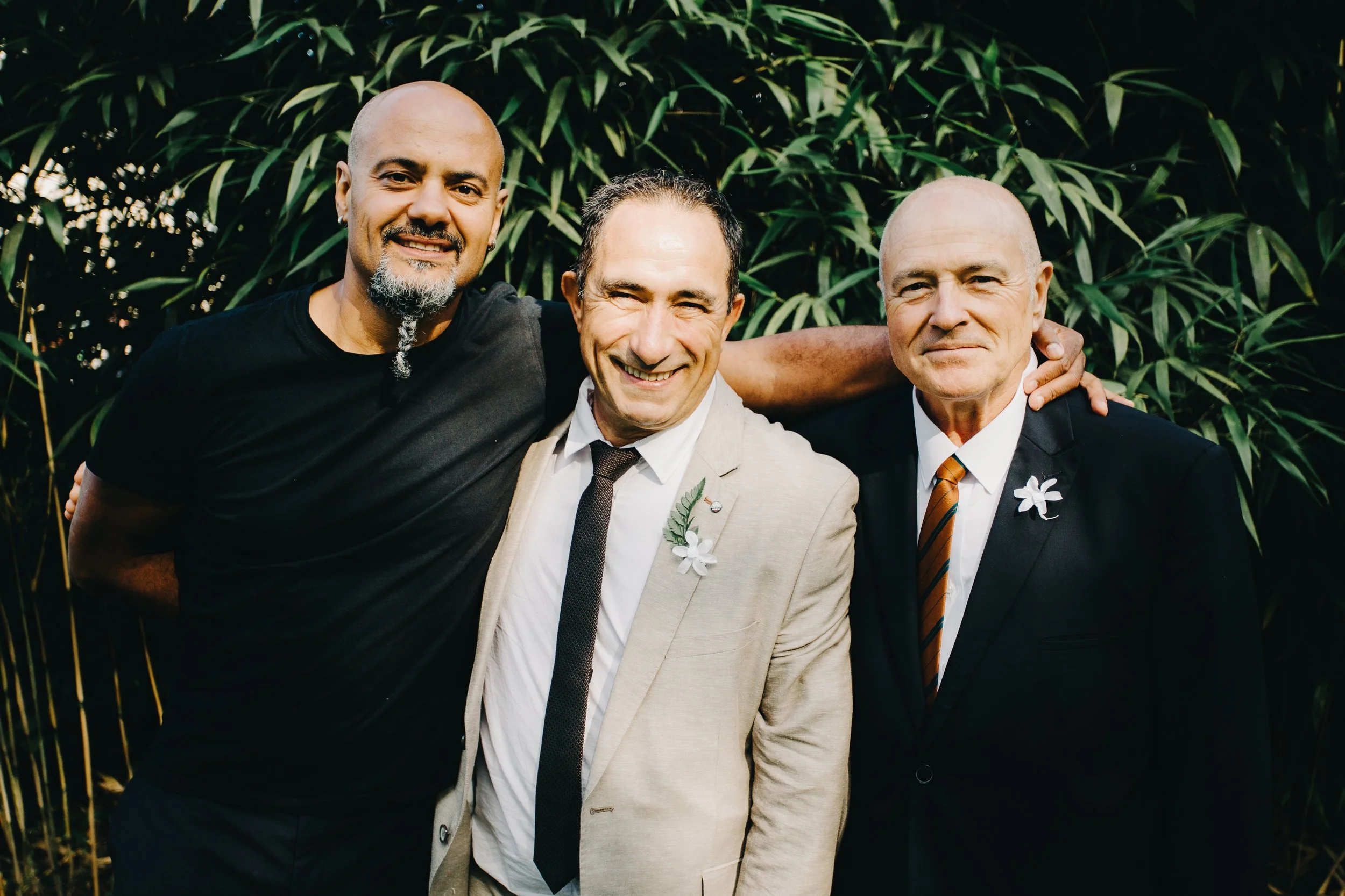 Trois hommes souriants portant des costumes, posant ensemble à l'extérieur devant un mur de feuilles vertes.
