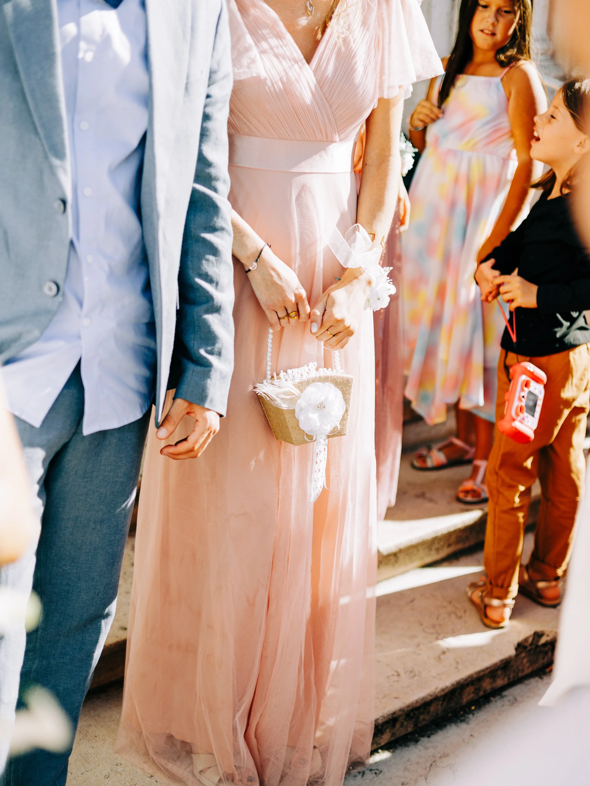 Groupe de personnes, dont un adulte en robe rose longue et des enfants, lors d'une célébration ou d'un événement en plein air, près d'une marche en pierre.