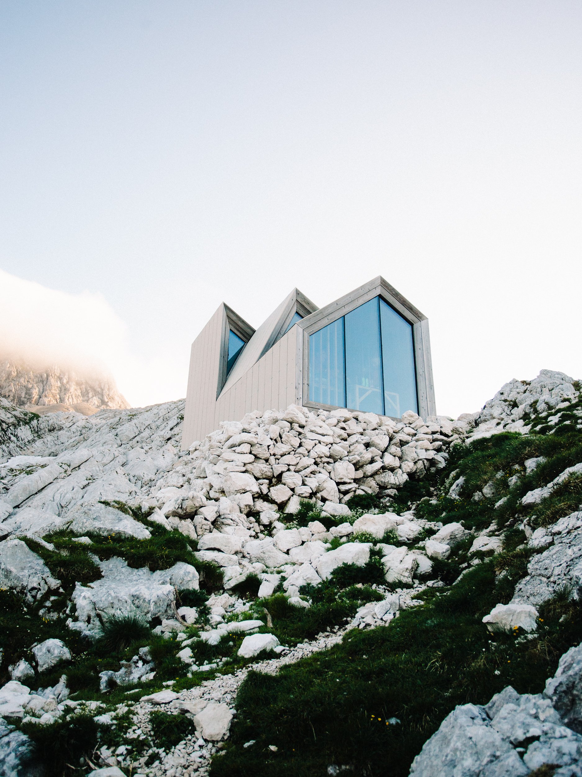 Montagne rocheuse avec une maison moderne en verre et en acier, située sur un terrain rocheux avec peu de végétation, sous un ciel clair.