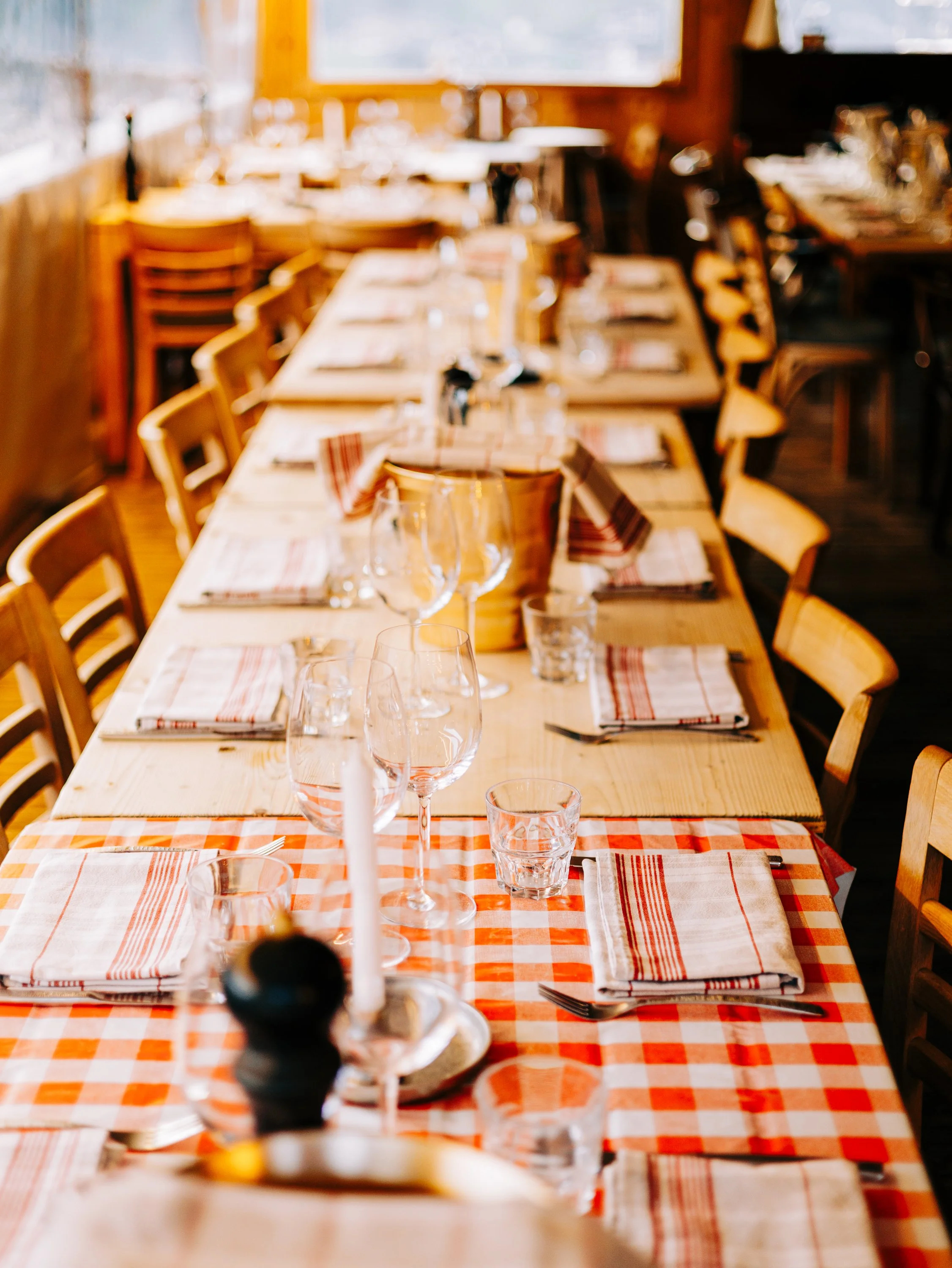 Une table dressée avec des verres, des assiettes, des couverts, et des serviettes en tissu à carreaux orange et blanc, dans une salle à manger chaleureuse avec beaucoup de lumière naturelle.