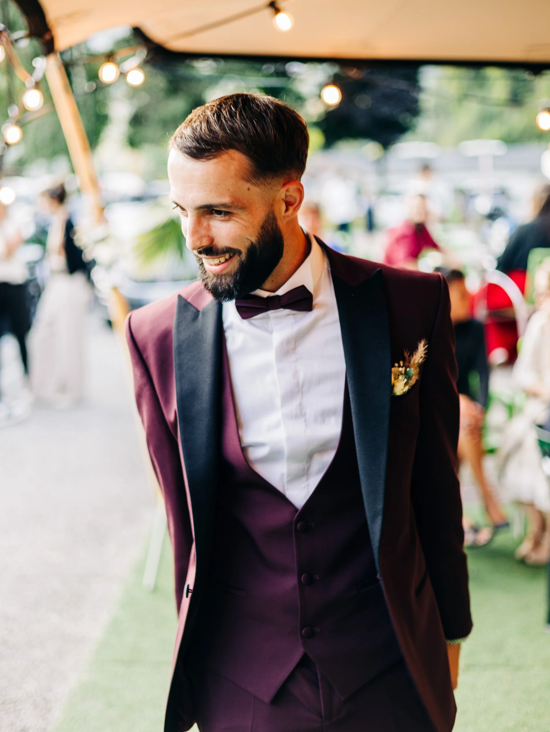 Un homme souriant portant un costume de mariage avec une veste bordeaux, une chemise blanche et un nœud papillon, lors d'une fête en extérieur décorée de guirlandes lumineuses.