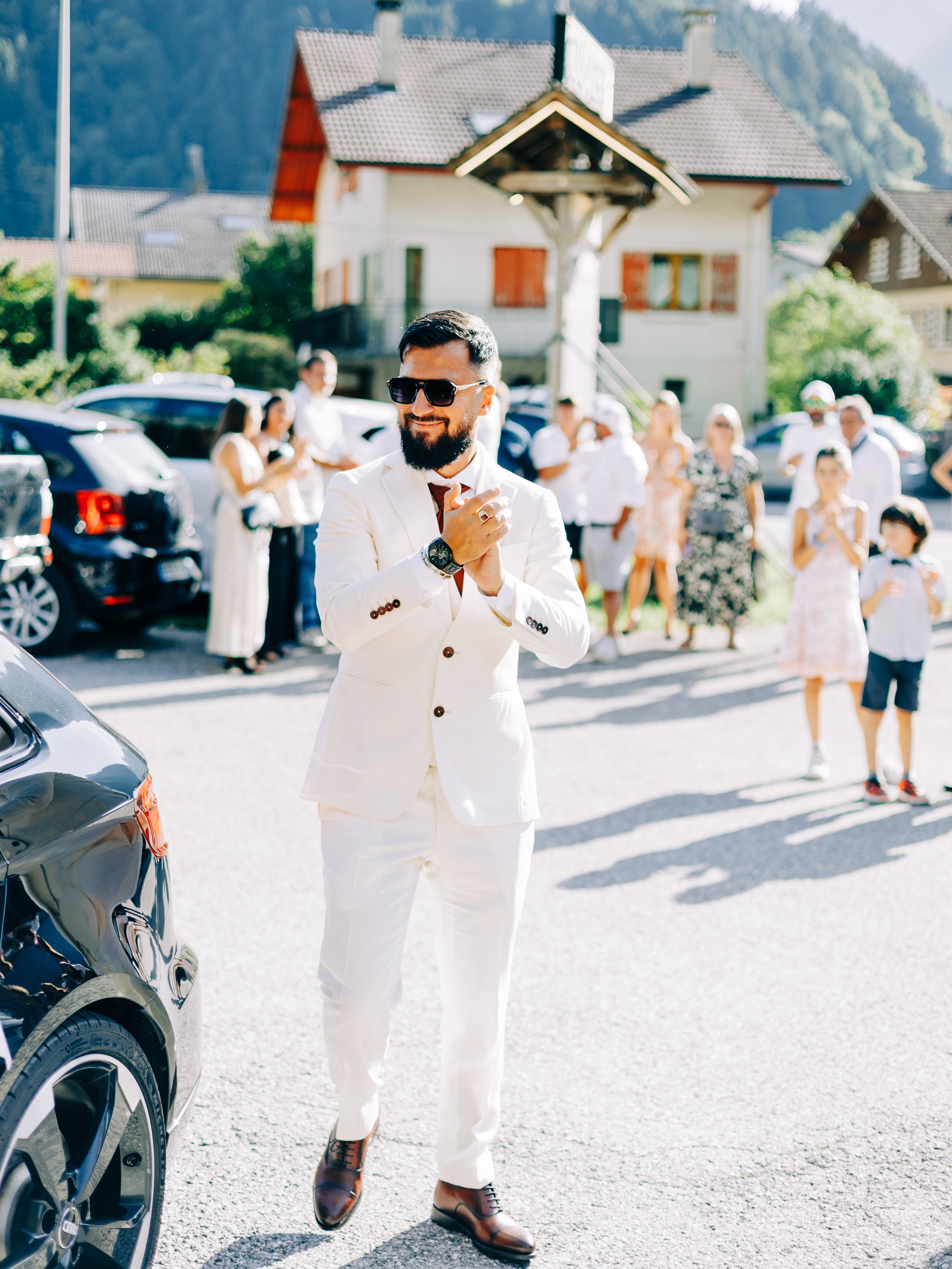 Un homme avec une barbe, portant des lunettes de soleil, un costume blanc, et des chaussures marron, sourit lors d'une fête en plein air avec d'autres personnes en arrière-plan, dans un village alpin avec des maisons et des voitures.