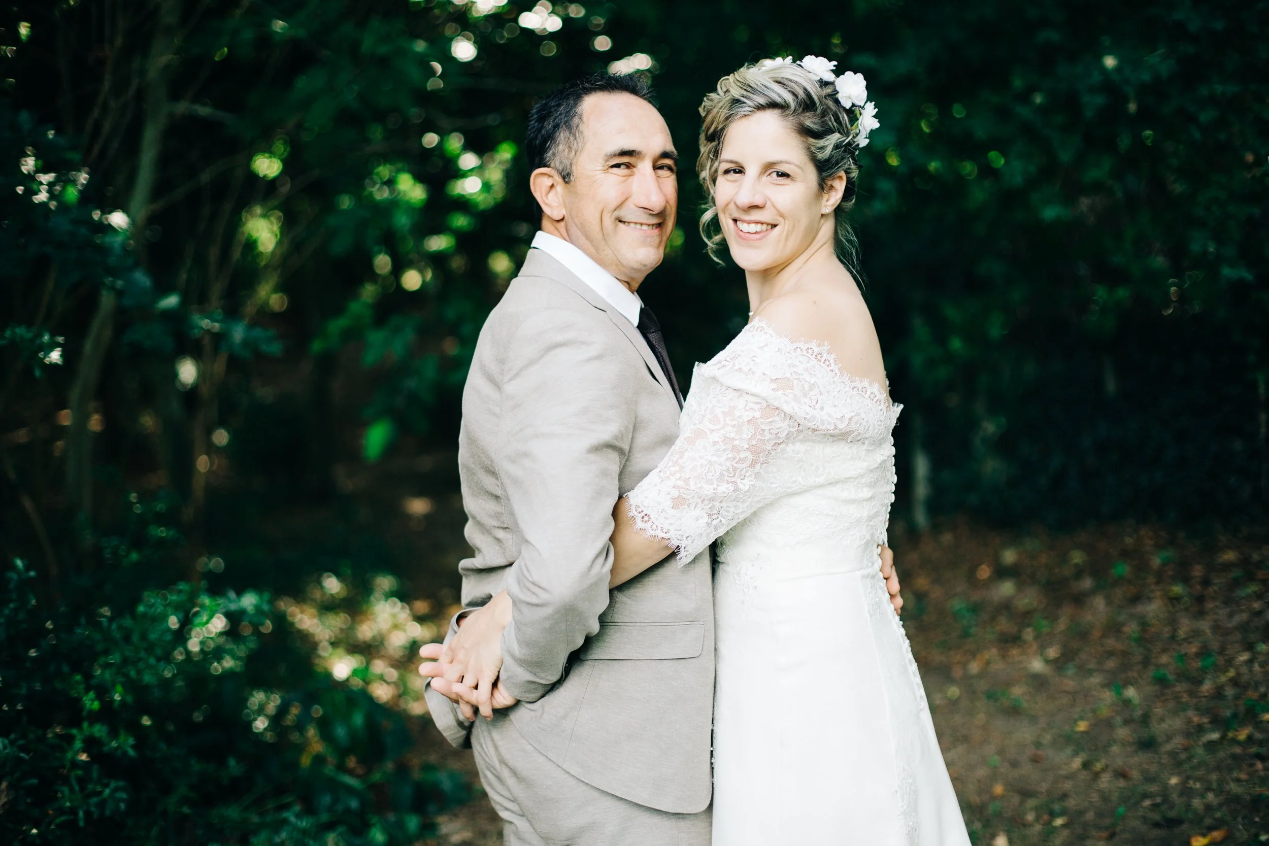 Un couple en mariage pose dans un jardin verdoyant, la femme porte une robe blanche avec des fleurs dans les cheveux, l'homme porte un costume beige.
