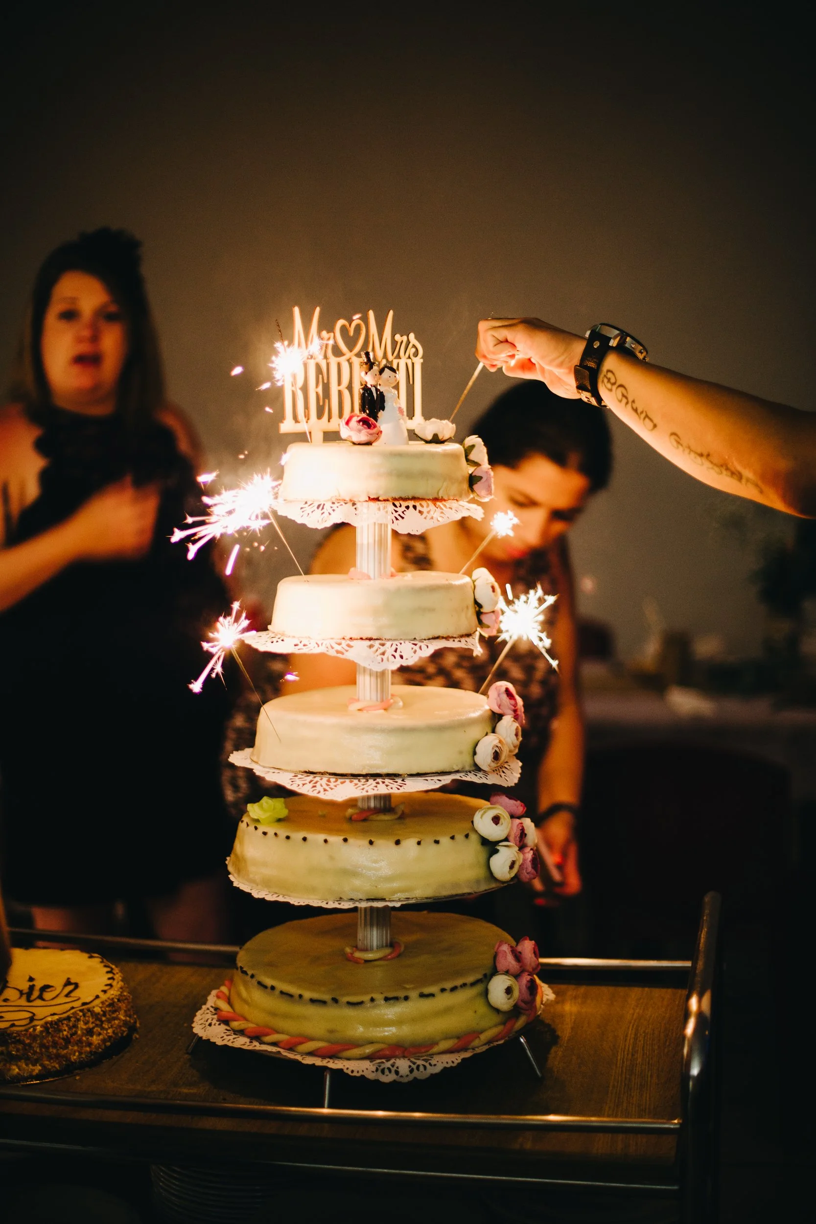 Gâteau de mariage à plusieurs étages avec des décorations florales et des gourmandises roses et blanches, avec des bougies et des lettres dorées pour "M + Mrs Rebell" sur le dessus, entouré de personnes célébrant une occasion spéciale.