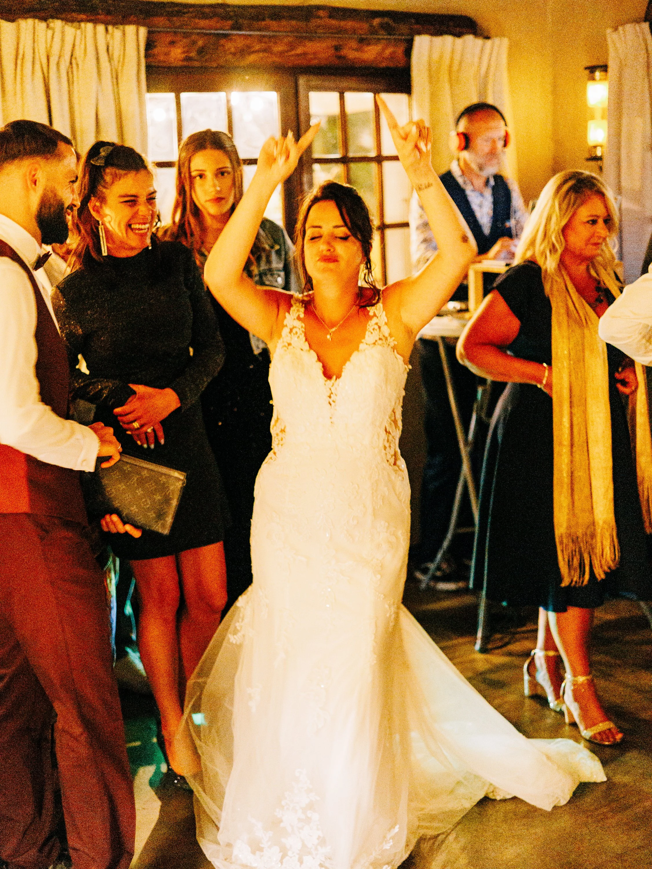 Une femme en robe de mariée blanc levant les bras dans une pièce chaleureuse lors d'une célébration ou d'un mariage, entourée de personnes souriantes.