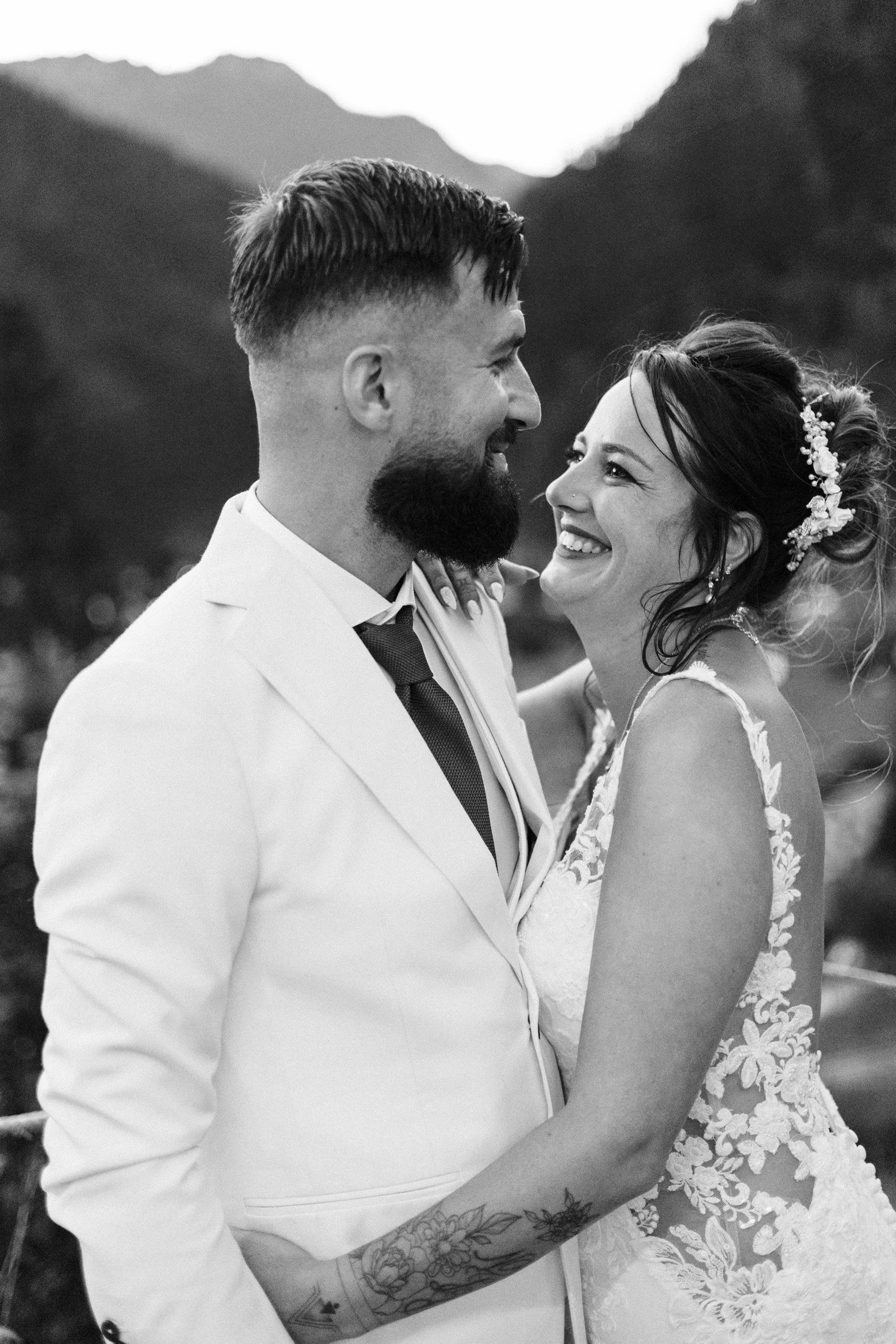 Un couple en mariage, homme en costume blanc avec barbe et tatouages, femme en robe de mariée, souriant, regardant dans les yeux, dans un environnement naturel montagnard.