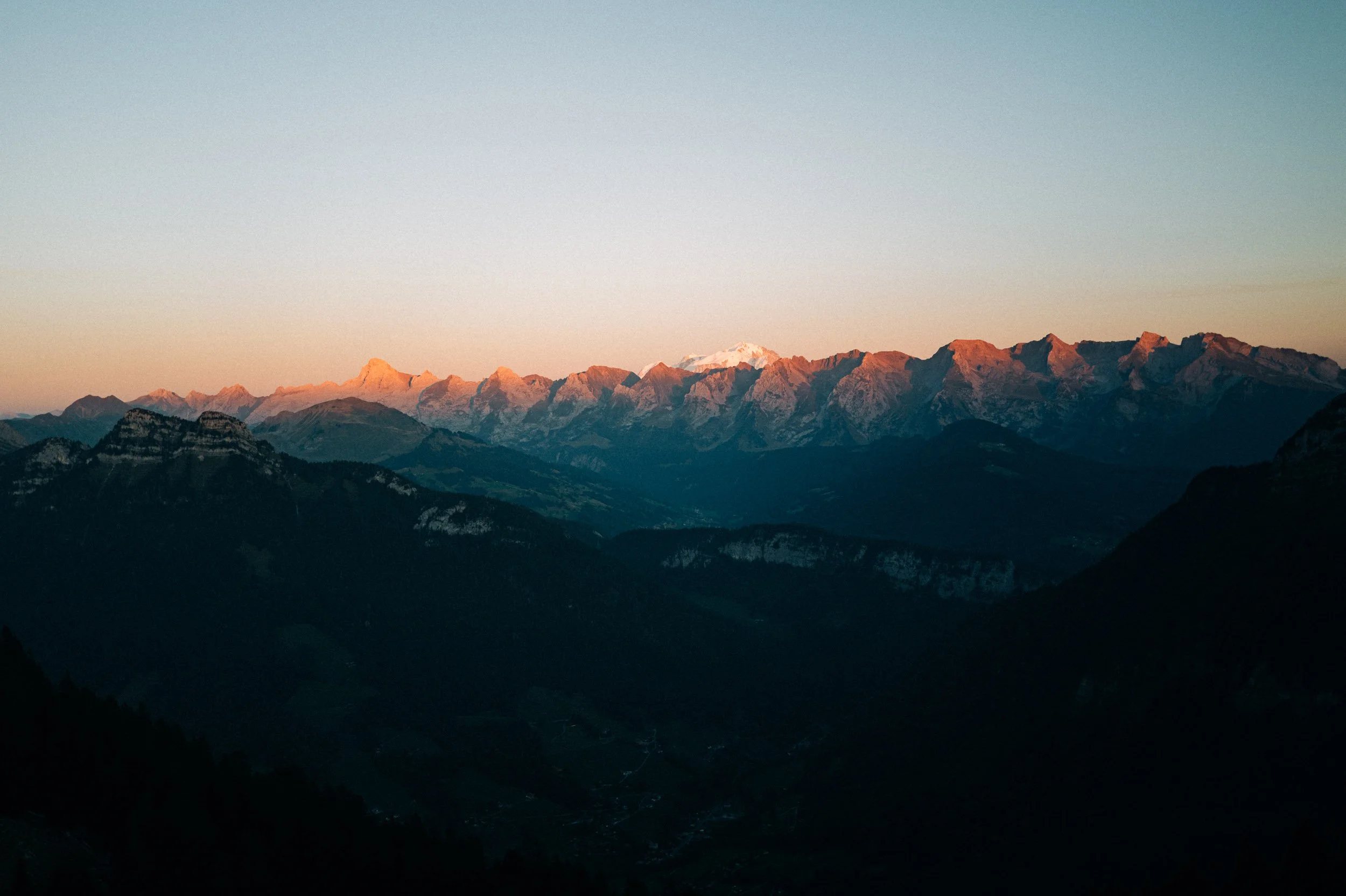 Vue d'une chaîne de montagnes au coucher du soleil, avec des sommets enneigés à l'horizon, dans un paysage naturel calme.