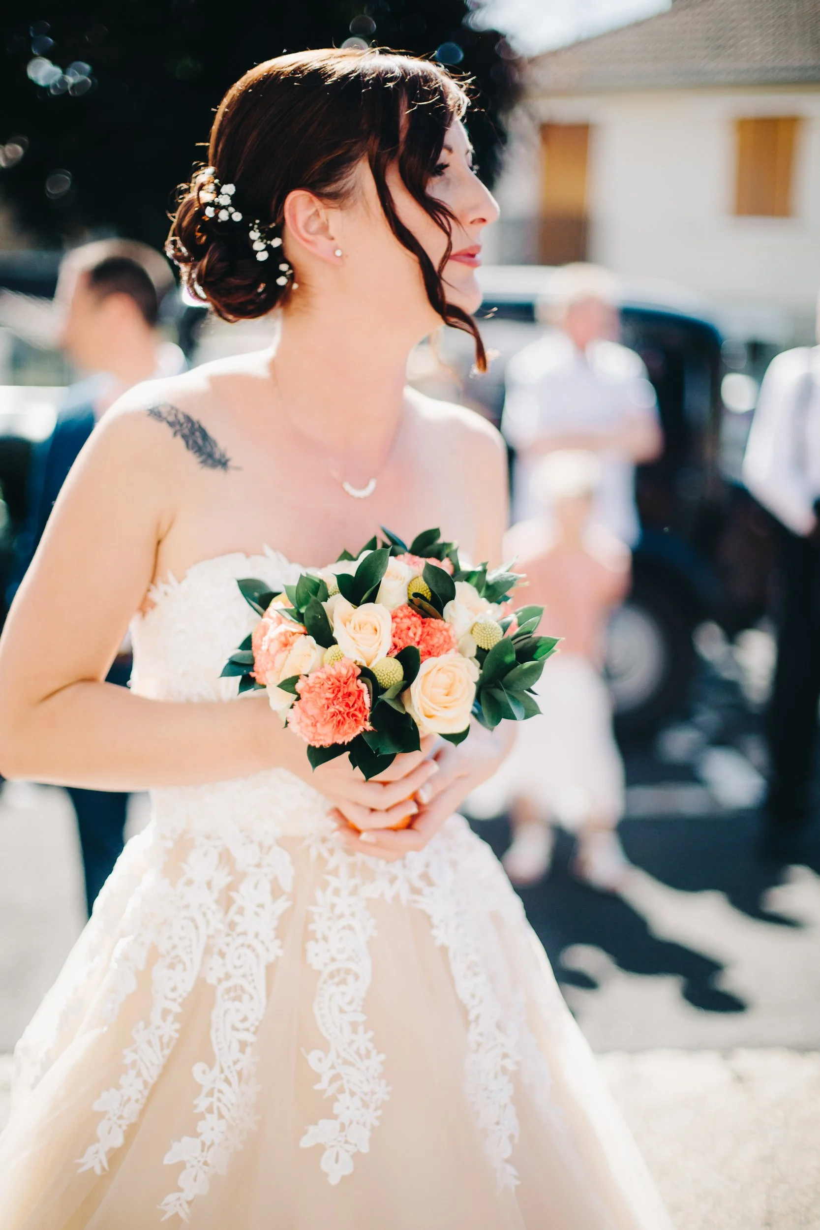Jeune femme en robe de mariage tenant un bouquet de fleurs, vue de profil, lors d'une cérémonie extérieure.