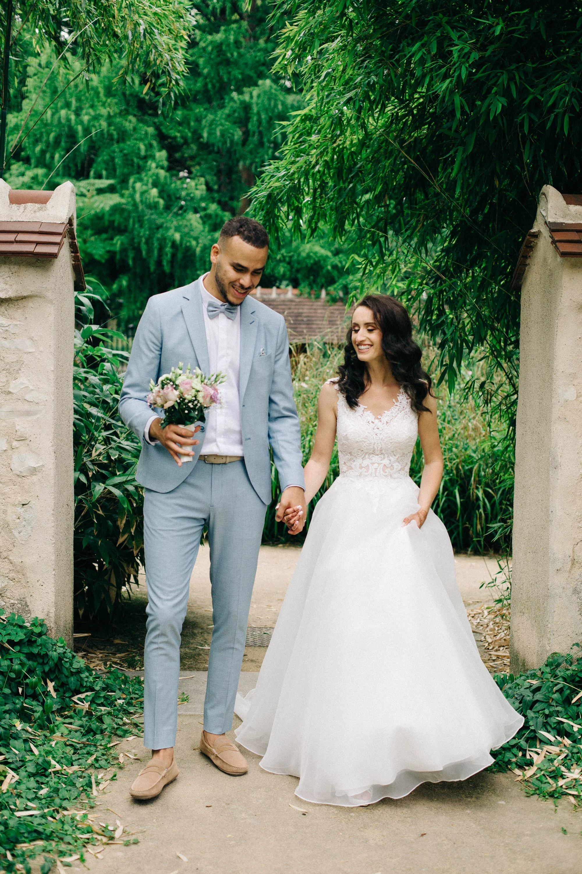 Un couple marié se tient sous une arche dans un jardin, mariée en robe blanche et le marié en costume gris clair, ils se tiennent par la main dans un cadre verdoyant.