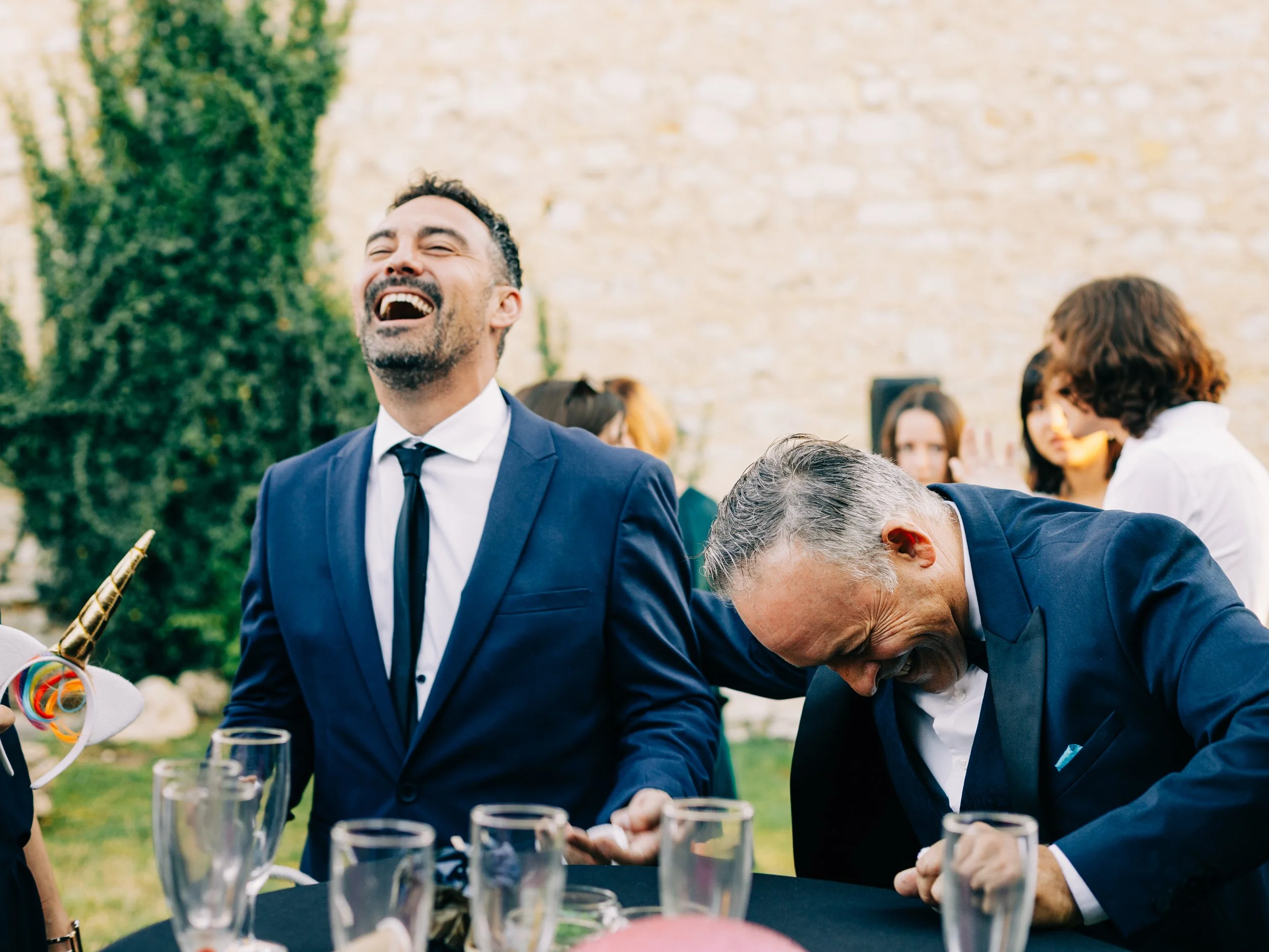 Groupe de personnes en costume lors d'une celebration en extérieur, lors d'un événement social ou professionnel, souriant et riant, avec une table décorée avec des verres et des décorations colorées.