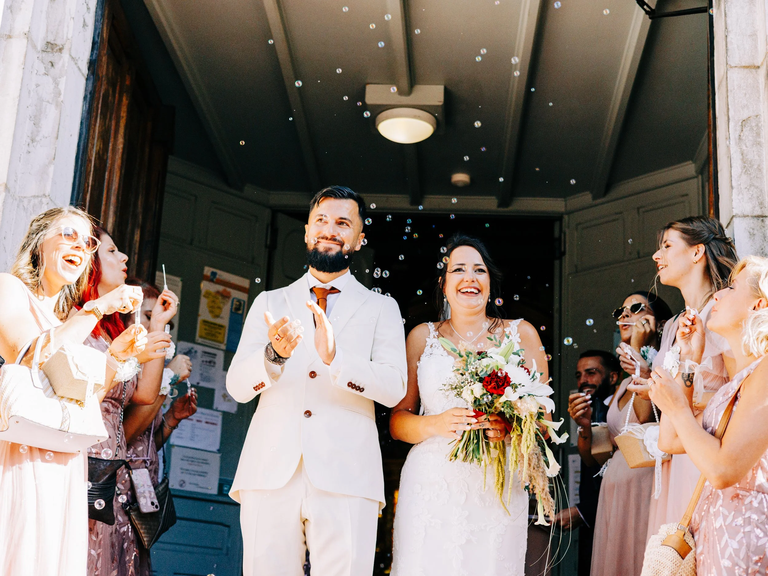 Un groupe de personnes lors d'un mariage, avec un couple au centre, la femme tenant un bouquet de fleurs et le homme portant un costume blanc, entourés d'invités célébrant, sous un ciel ensoleillé.