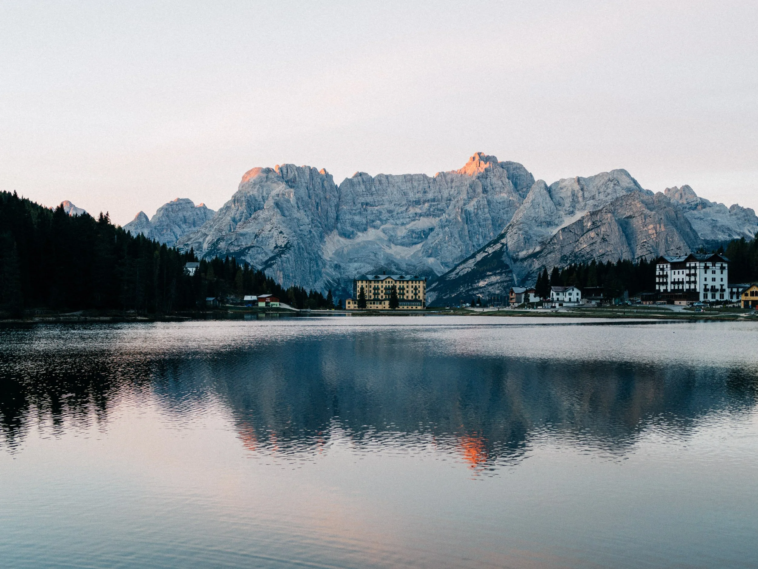 Montagnes rocheuses au coucher du soleil avec un lac au premier plan et un bâtiment en bas à droite.