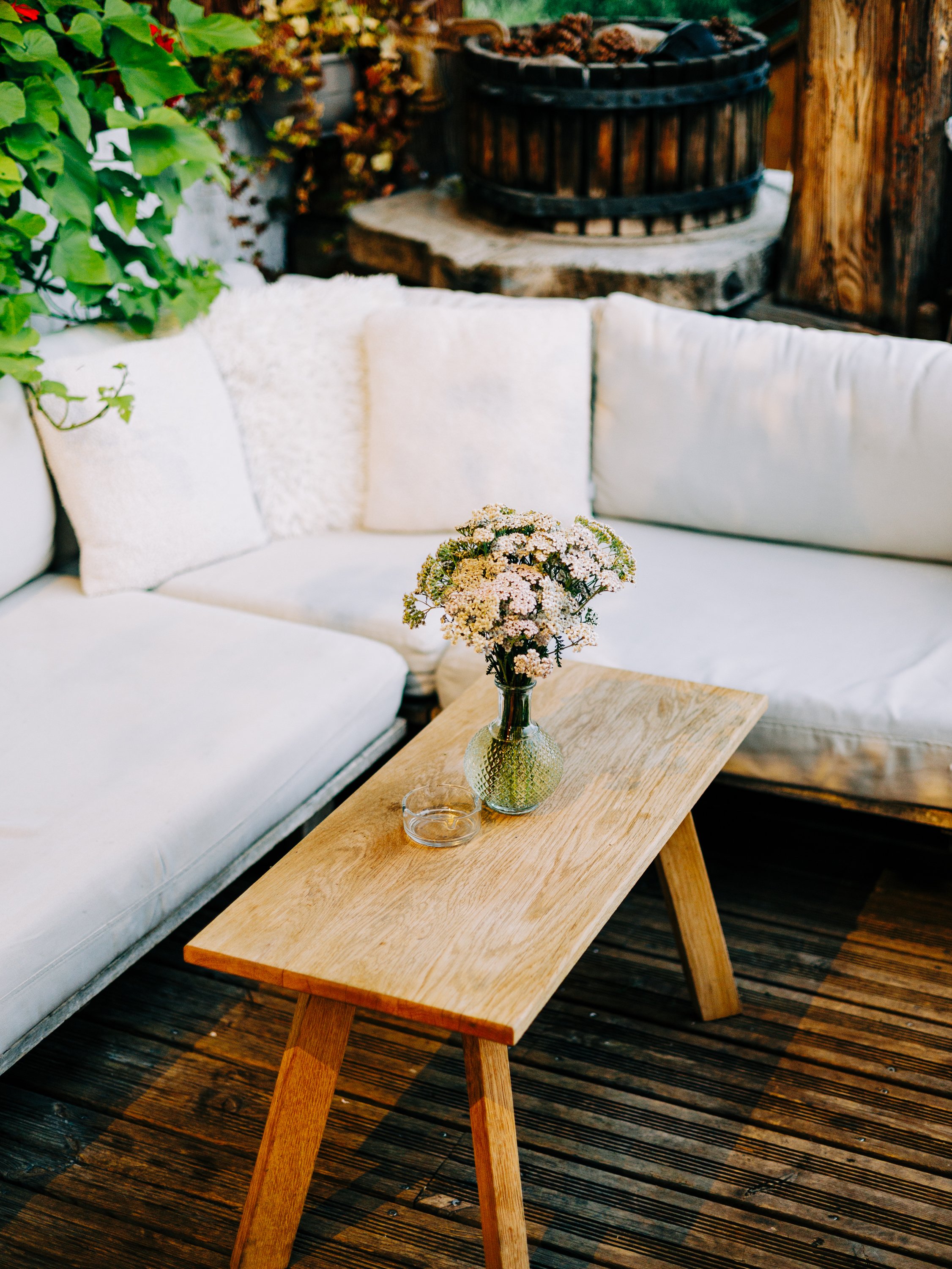 Table en bois avec une vase contenant des fleurs blanches et roses, un verre vide à côté, un canapé blanc avec deux coussins et un fond de décoration en bois avec plantes vertes