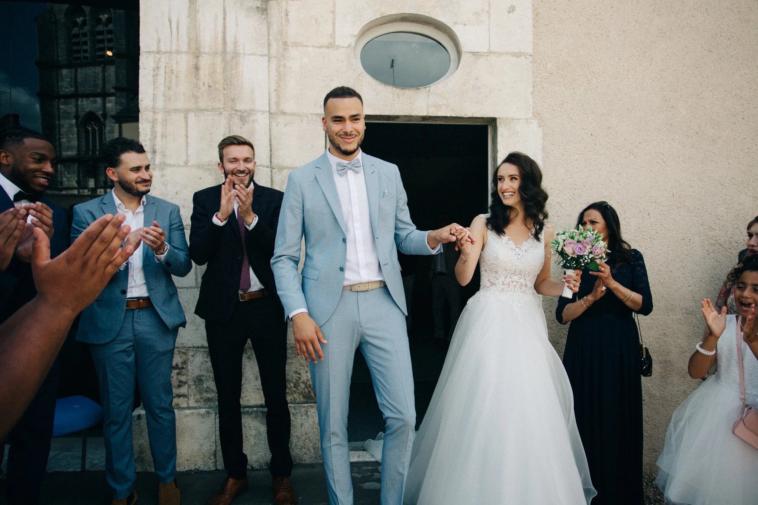 Un couple marié entouré de proches lors d'une cérémonie devant un bâtiment en pierre, tous souriant et applaudissant.
