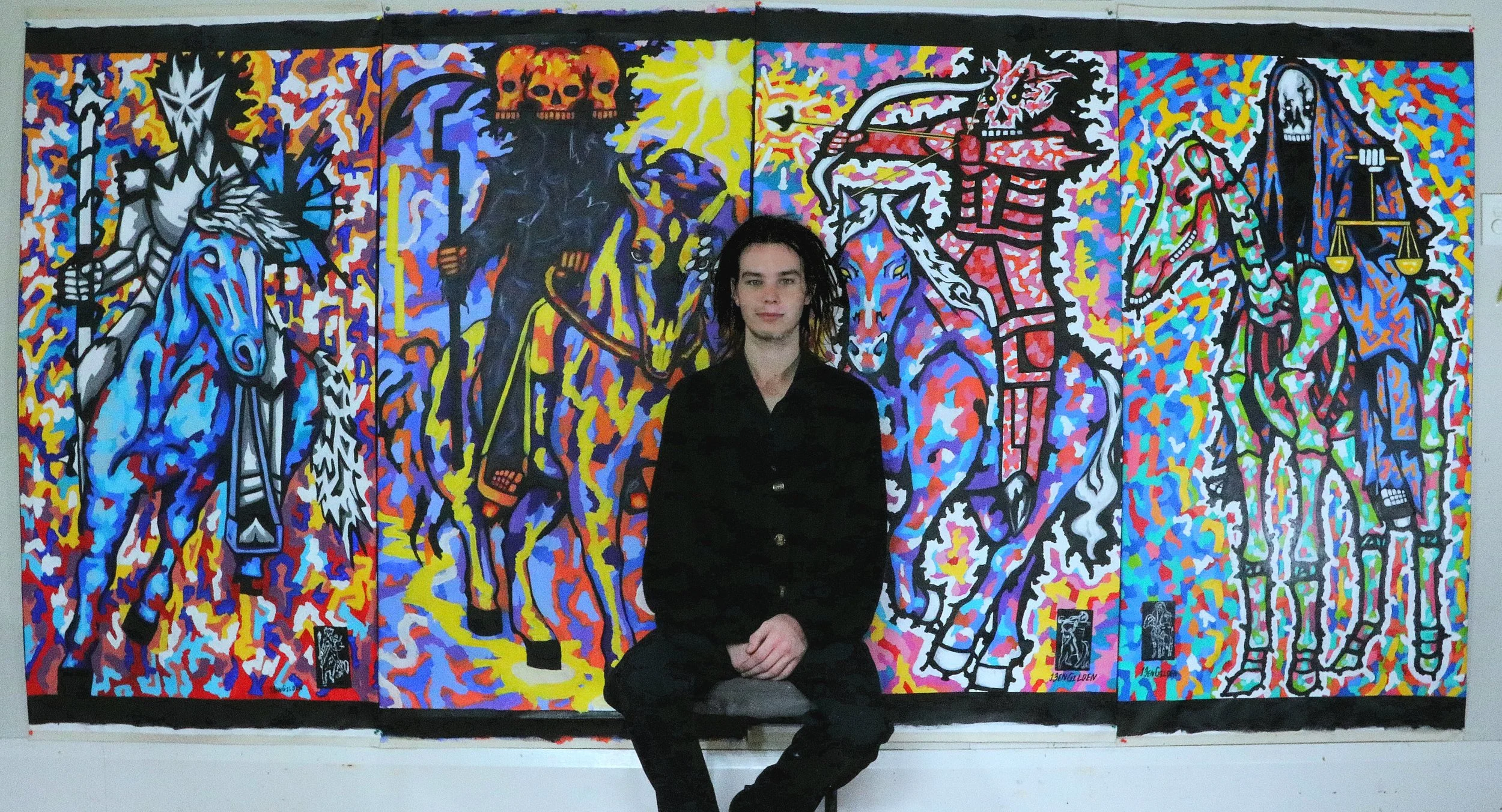 A person with long dark hair and black clothing sitting in front of a vibrant, colorful mural with abstract skeleton and creature illustrations.