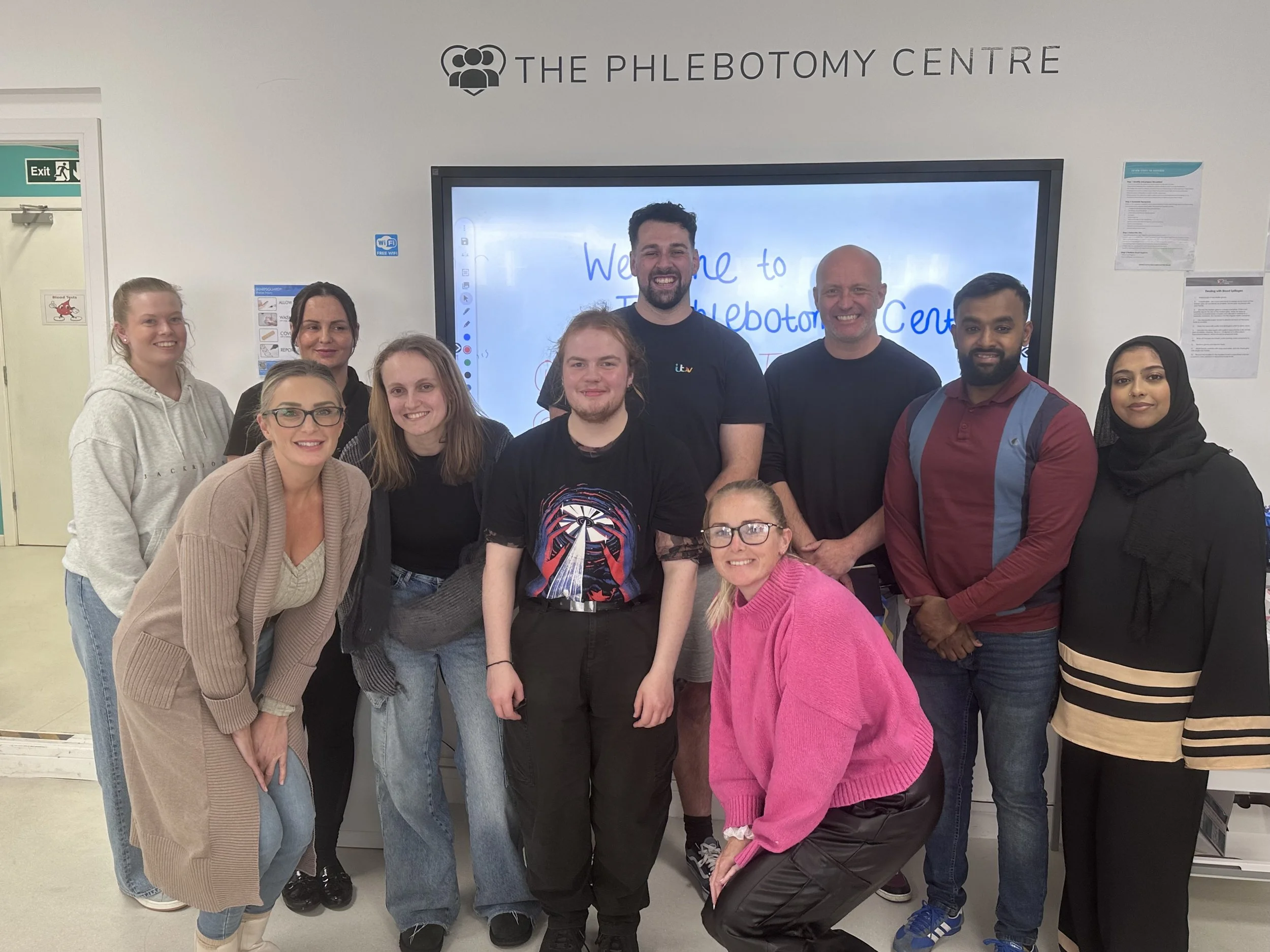 A group of ten people, including men and women, standing together indoors in front of a large screen displaying the words "Welcome to The Phlebotomy Centre." They are smiling and posing for the photo.