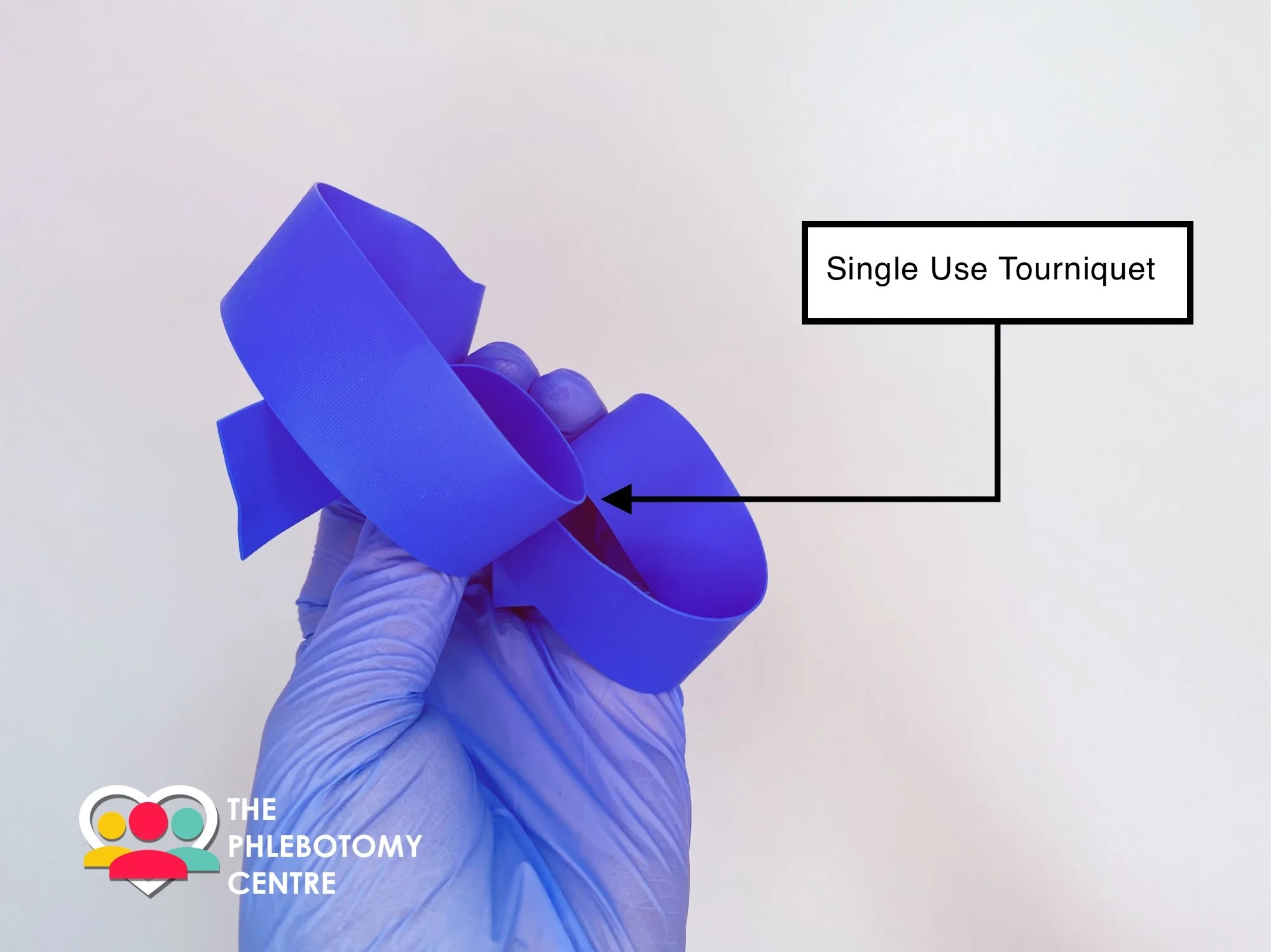 A gloved hand holding a blue single-use tourniquet against a plain white background, with a logo for The Phlebotomy Centre in the bottom left corner.