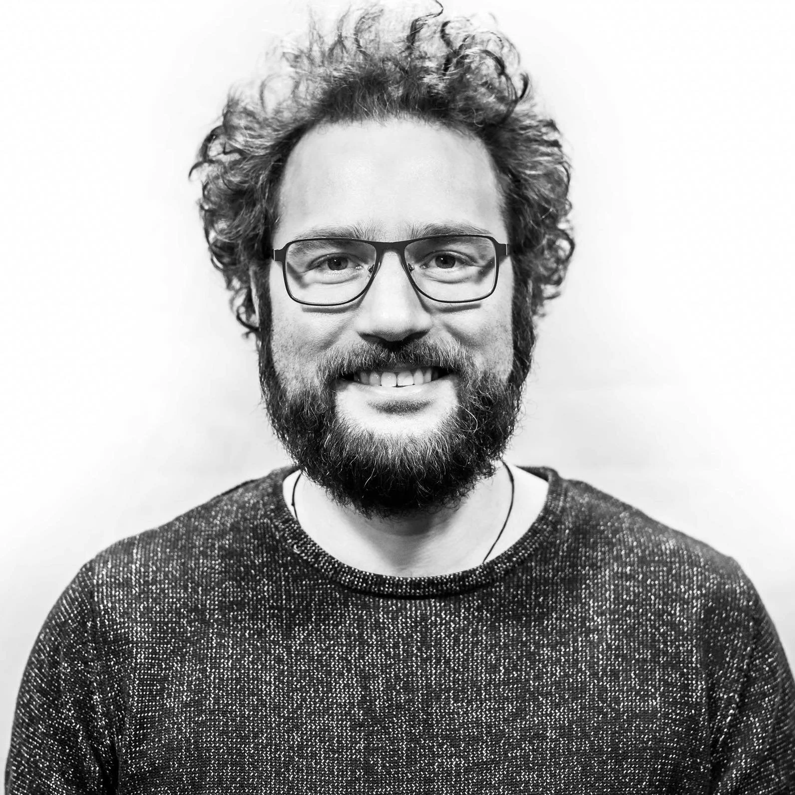 A black-and-white photo of a smiling man with curly hair, glasses, a beard, and wearing a textured sweater.