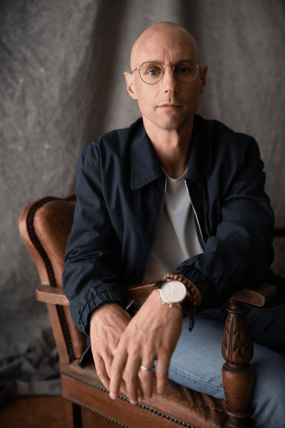 Life-writer and novelist Craig Shubin, with glasses sitting on a vintage wooden chair against a gray textured wall, wearing a dark jacket, white shirt, and jeans, with a watch and bracelets on his wrist.