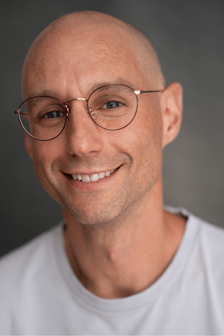 Close-up portrait of a life-writer and novelist Craig Shubin with glasses, wearing a white shirt, against a gray background.