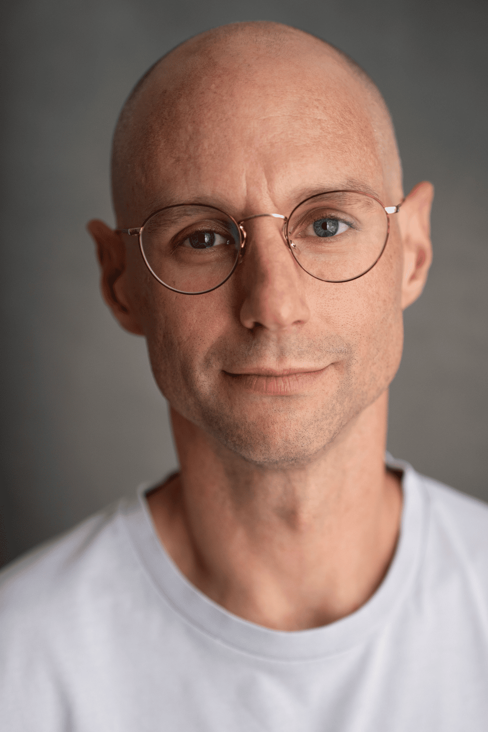 Close-up portrait of a life-writer and novelist Craig Shubin, wearing round glasses and a white t-shirt, smiling subtly against a gray background.