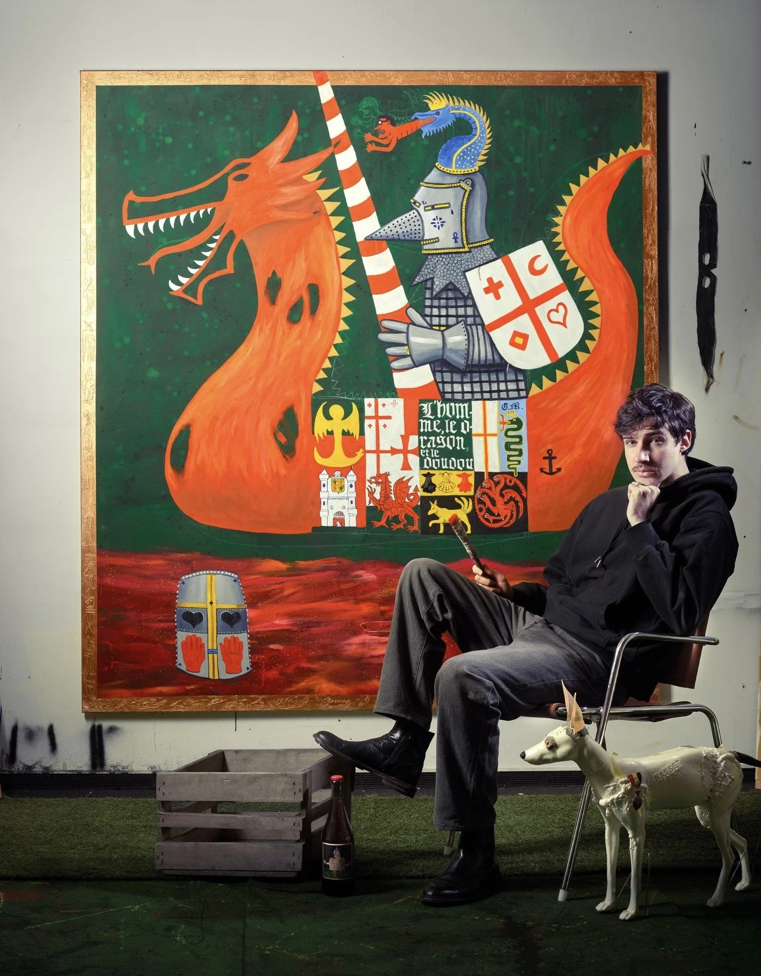 Un jeune homme assis dans un fauteuil, portant une veste noire et tenant une cigarette, à côté d'une statue de chien en plastique. Sur le mur en arrière, une peinture colorée représentant un chevalier en armure avec un bouclier, un dragon et d'autres motifs symboliques.