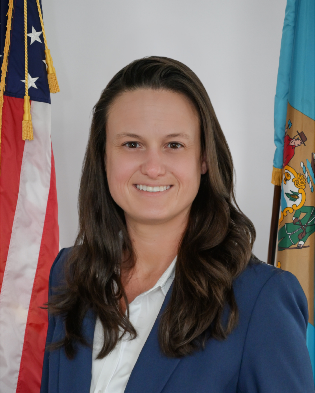 Tori Parker in a blue blazer and white shirt standing in front of the American and State of Delaware flags.
