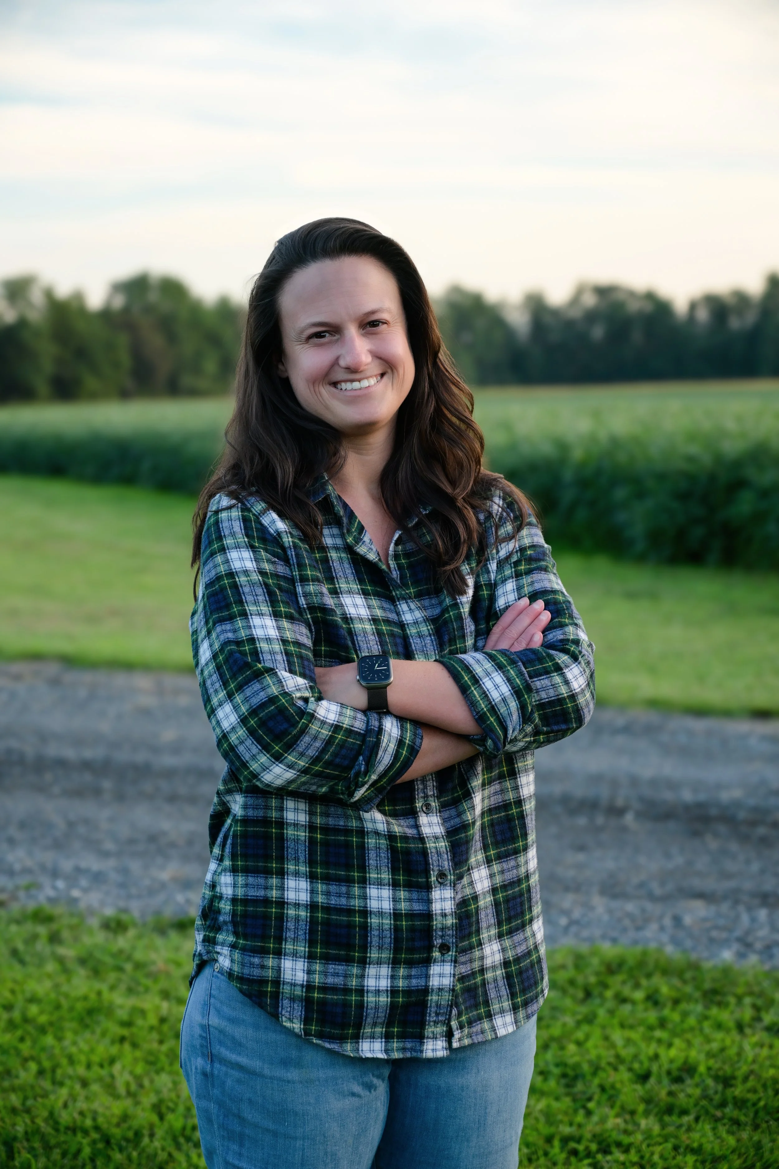 Tori Parker pictured with long dark hair smiling outdoors, wearing a plaid shirt and a smartwatch, with her arms crossed, in front of a field and trees under a cloudy sky.
