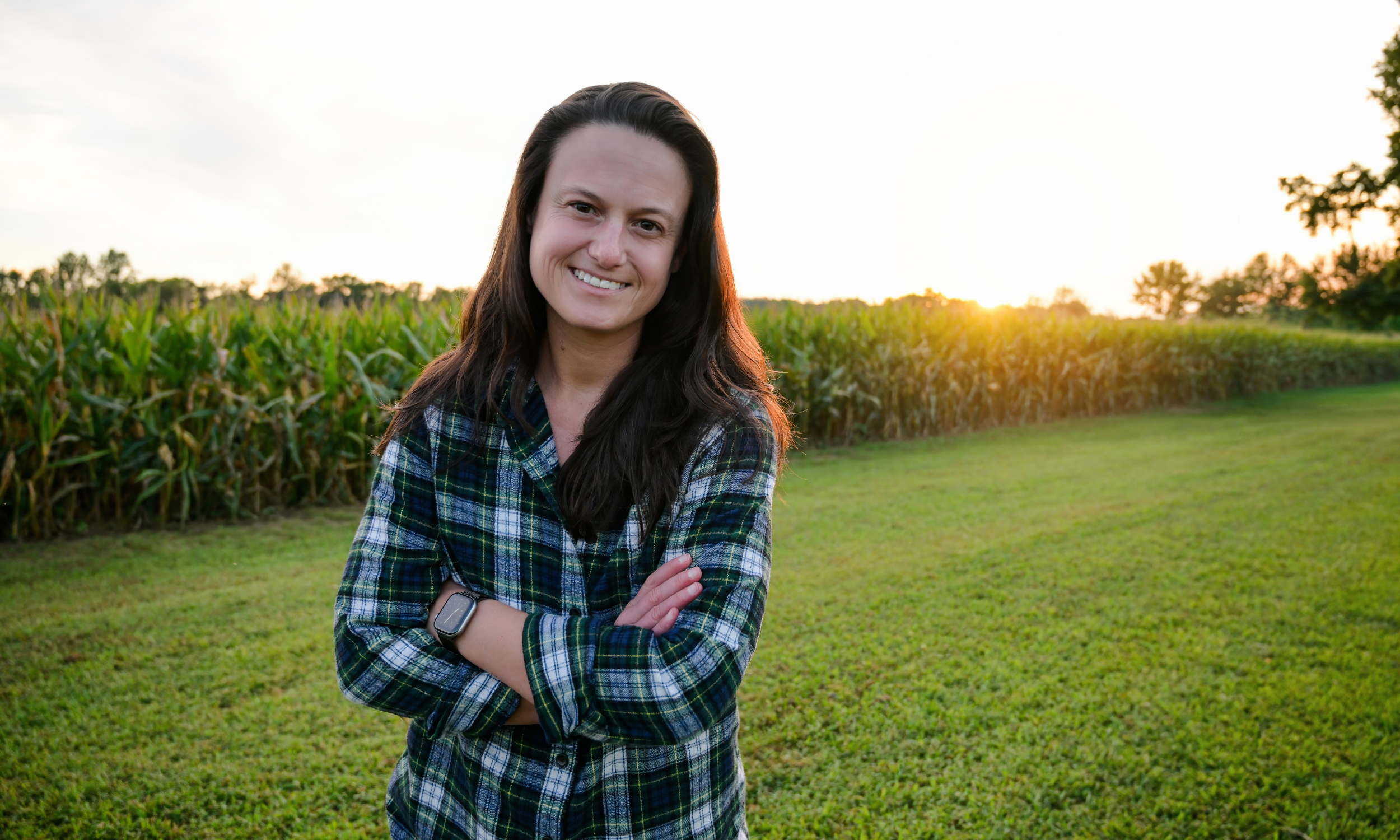 Tori Parker.  long dark hair, wearing a green plaid shirt and a smartwatch, standing outdoors in front of a green field with corn plants at sunset.