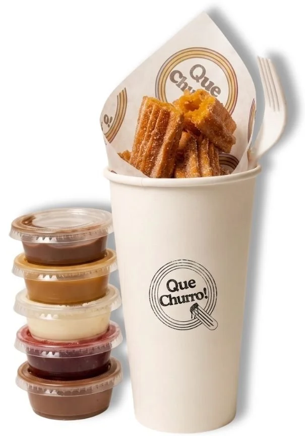 A white paper cup with the Que Churro! logo holding churros, with small containers of different customized flavors of dipping sauces stacked next to it.