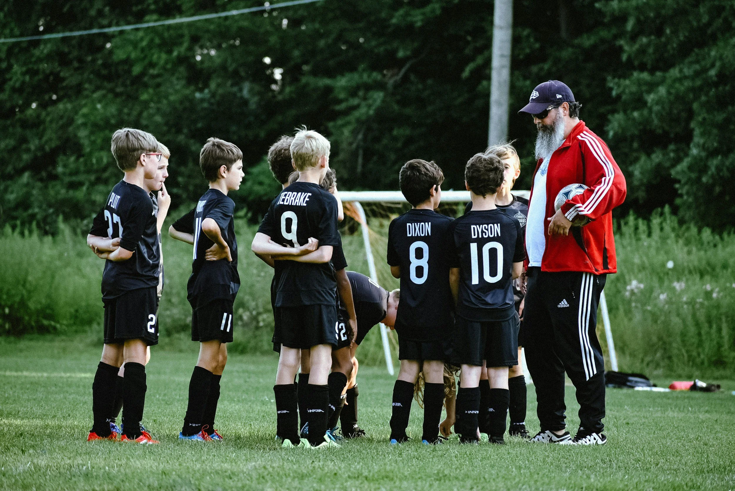 A coach in a red jacket and black cap with sunglasses talking to young soccer players on a field.