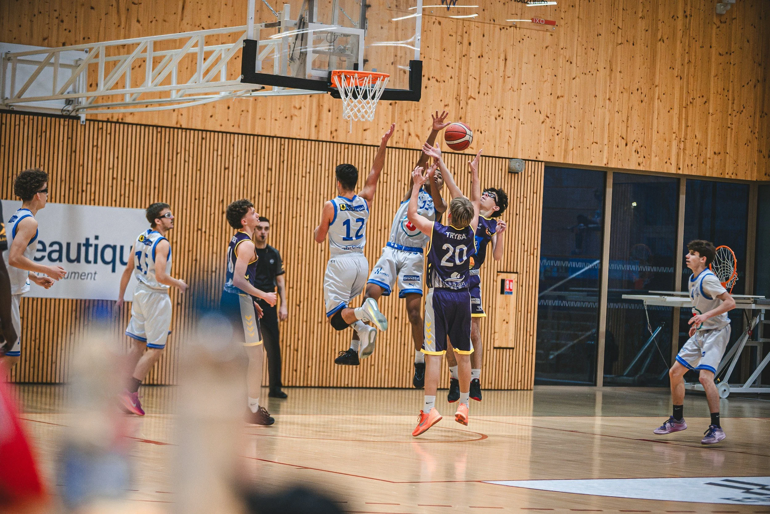 Youth basketball game inside gym, players jump to catch ball near basket, wooden wall background, players in white, blue, yellow, and purple uniforms.