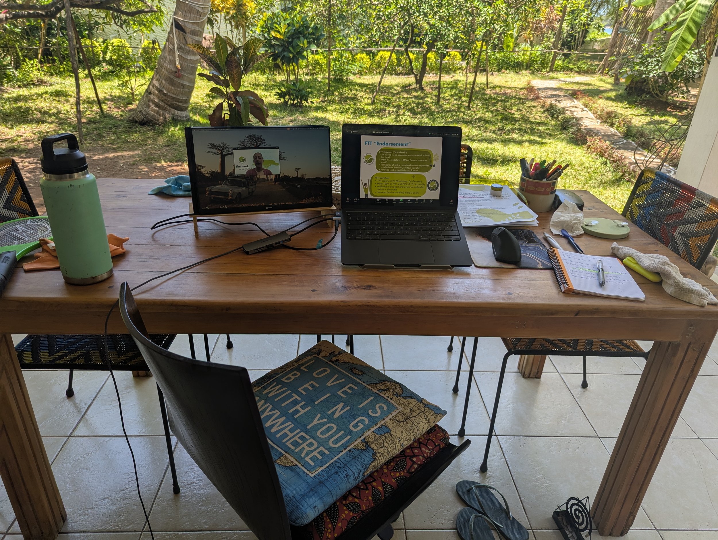 Table en bois avec un ordinateur portable, un écran d'ordinateur, un carnet, des stylos, une gourde, une tasse, des documents et d'autres objets, avec un jardin verdoyant en arrière-plan.