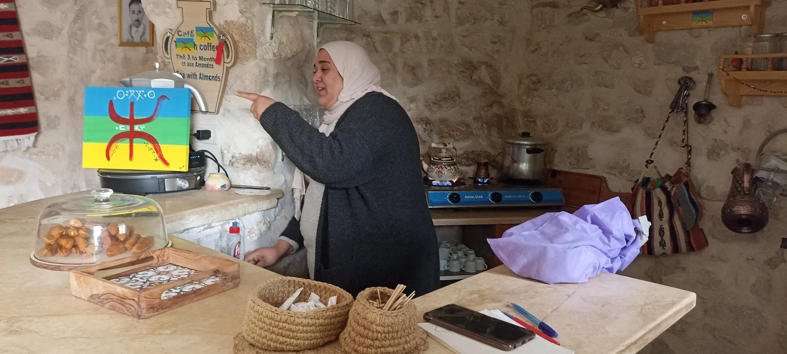 Une femme portant un foulard blanc et un vêtement grisaille pointant du doigt vers la gauche dans une cuisine rustique en pierre. Sur le comptoir, il y a une boîte en bois contenant des biscuits, un robot de cuisine, deux paniers en osier, un téléphone, et des objets de cuisine. Le fond montre une cuisinière à gaz, des étagères avec des pots, une lampe suspendue et un tableau avec un dessin coloré.