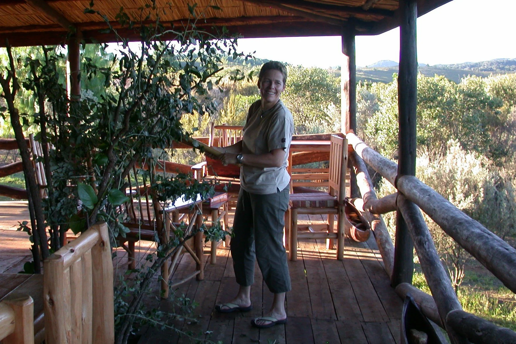 Une femme souriante debout sur une terrasse en bois entourée de végétation, tenant un livre, avec un paysage de collines et de ciel clair en arrière-plan.