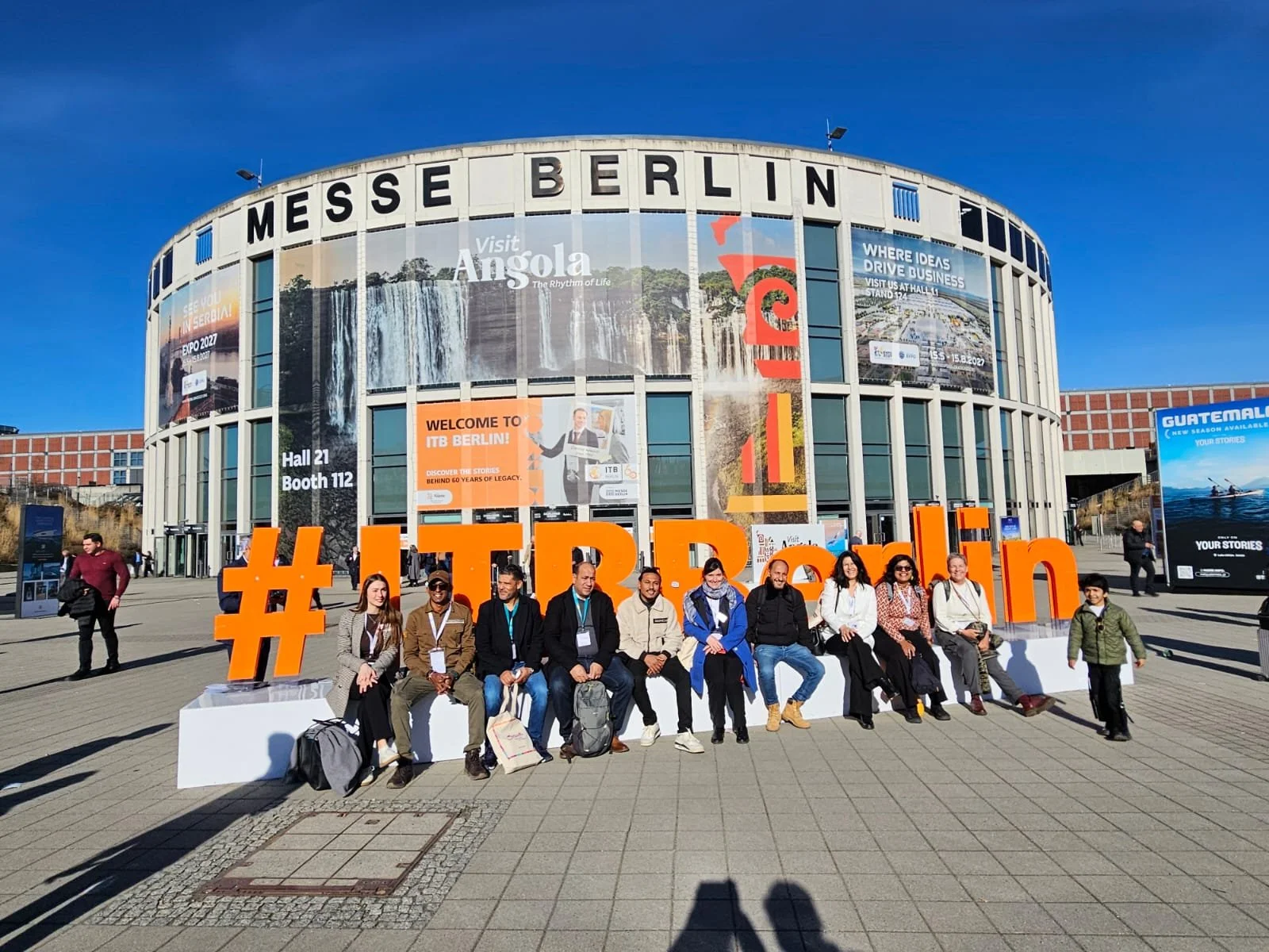 Groupe de personnes assises devant l'entrée du centre d'exposition Messe Berlin, avec un grand symbole #ITRBerlin en orange devant elles.