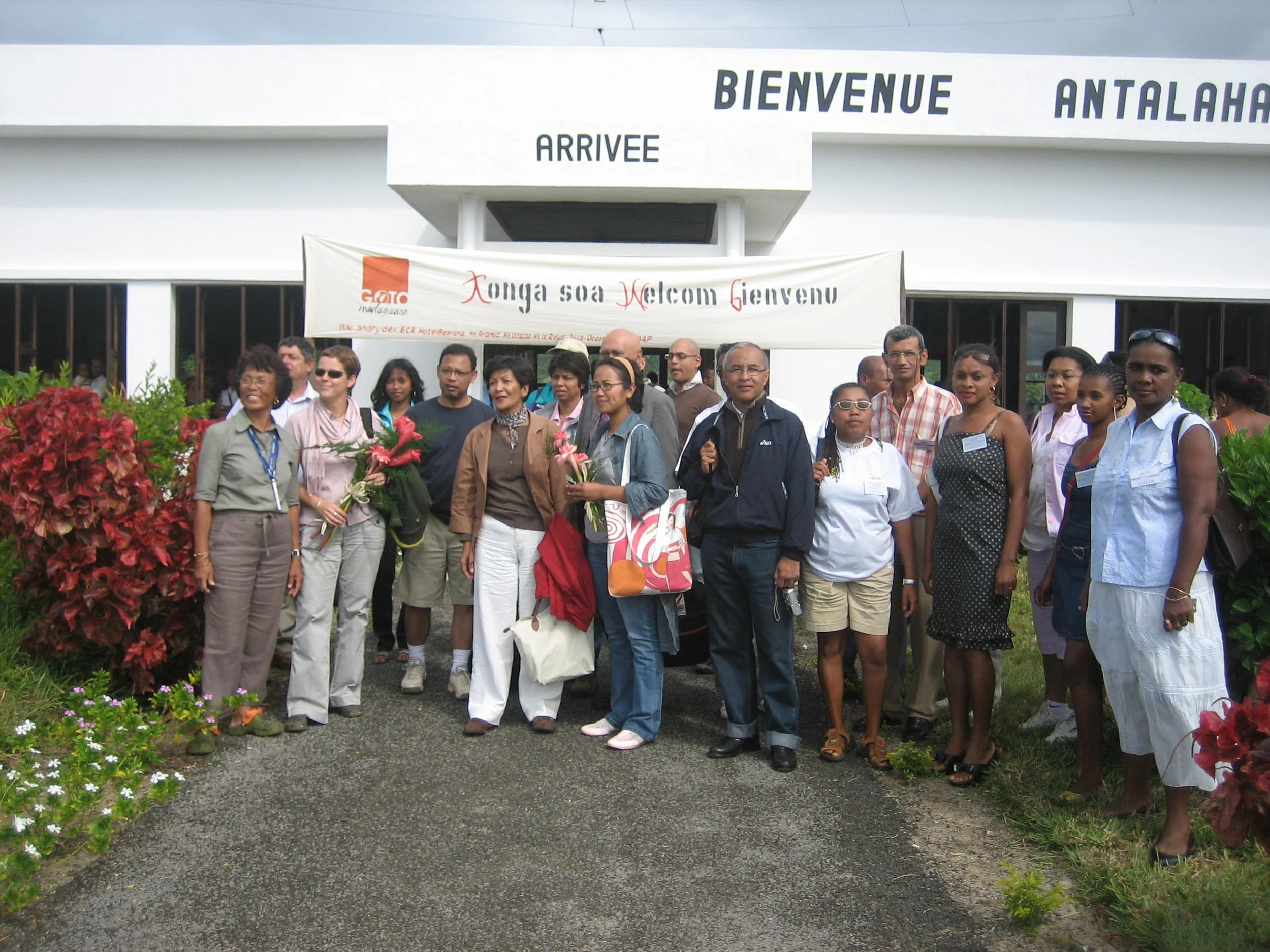 Groupe de personnes devant l'entrée de l'aéroport d'Antalaha, avec un panneau disant 'Bienvenue Antalaha' et portant des fleurs, semble accueillir une délégation ou un groupe de visiteurs