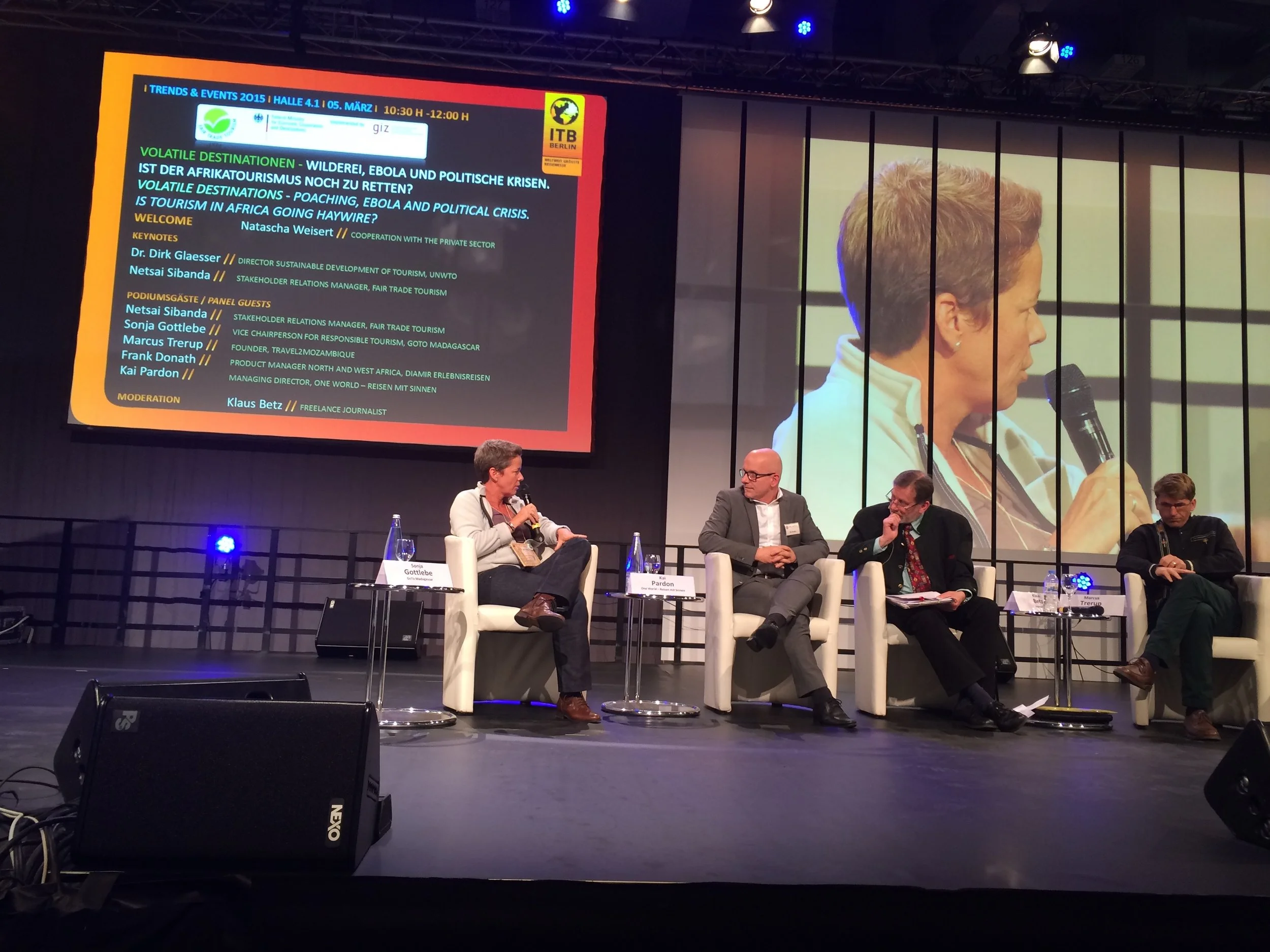 Une conférence ou un panel avec plusieurs intervenants assis sur scène, un écran en arrière-plan affiche un programme en allemand, anglais et français sur le tourisme en Afrique, la crise politique et le virus Ebola, ainsi que les noms des intervenants et leurs titres.