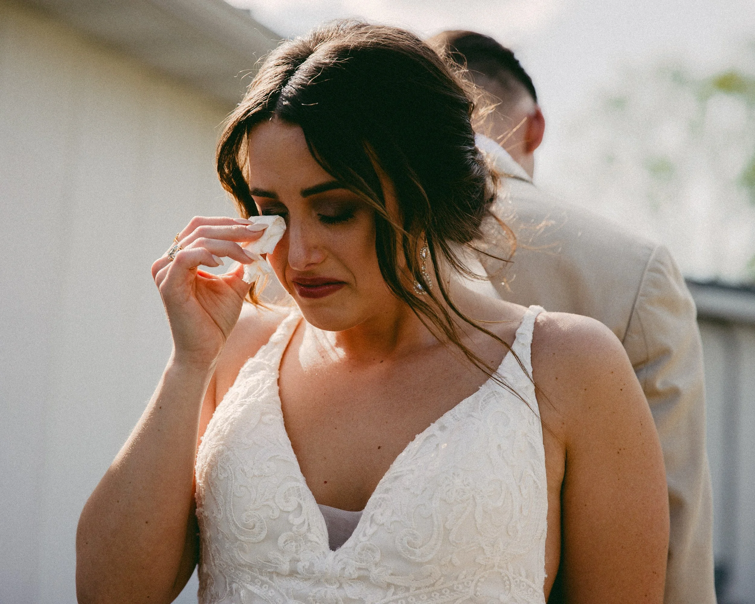 A woman in a white lace dress, likely at her wedding, is crying and wiping tears from her face with tissue. She is standing close to a man in a light-colored suit. St. Louis Documentary-style wedding photography on 35mm film, 120 film, and digital by