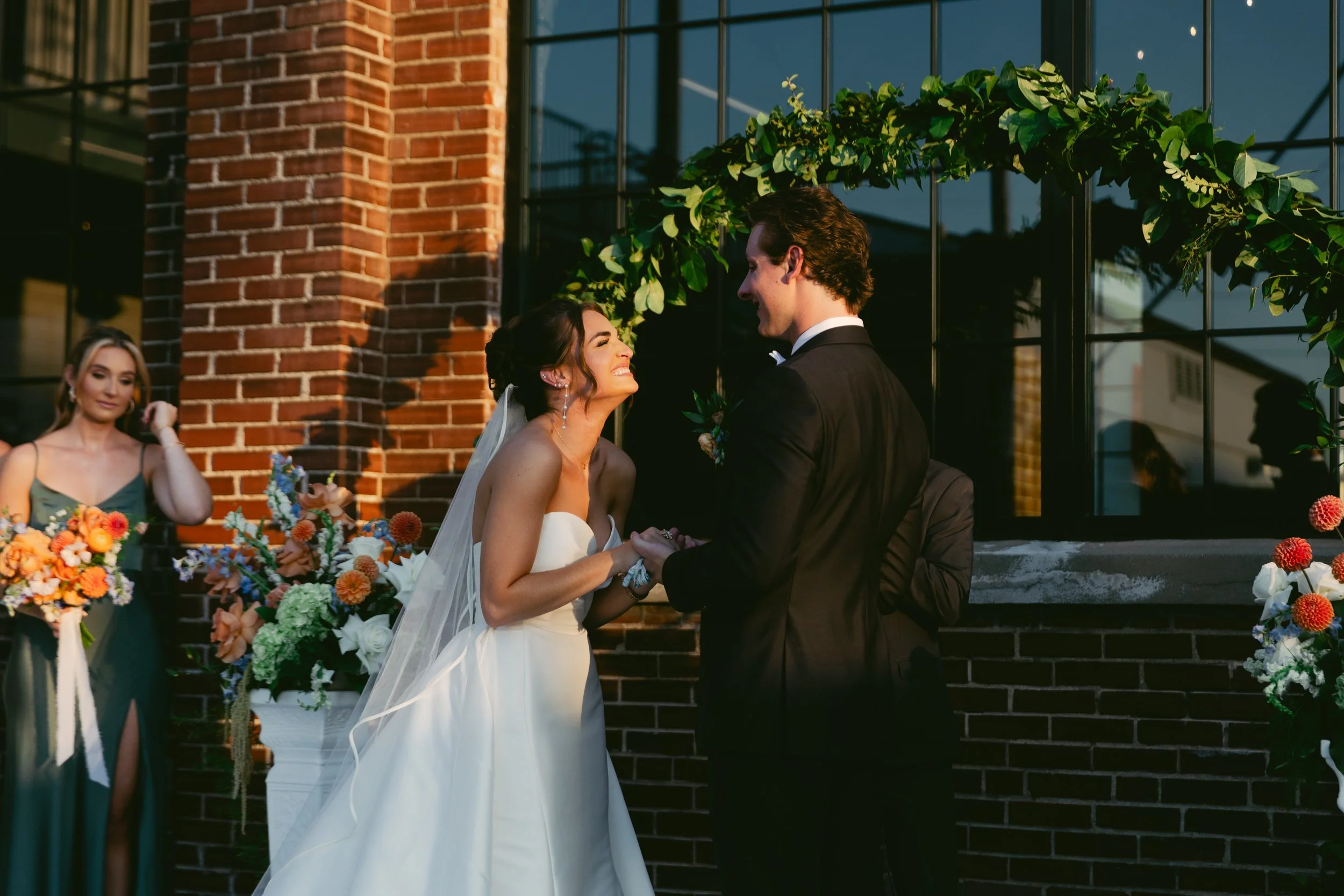 A bride and groom holding hands and smiling at each other during their wedding ceremony, with bridesmaids and floral arrangements outside a brick building. St. Louis Documentary-style wedding photography on 35mm film, 120 film, and digital by Trevor 