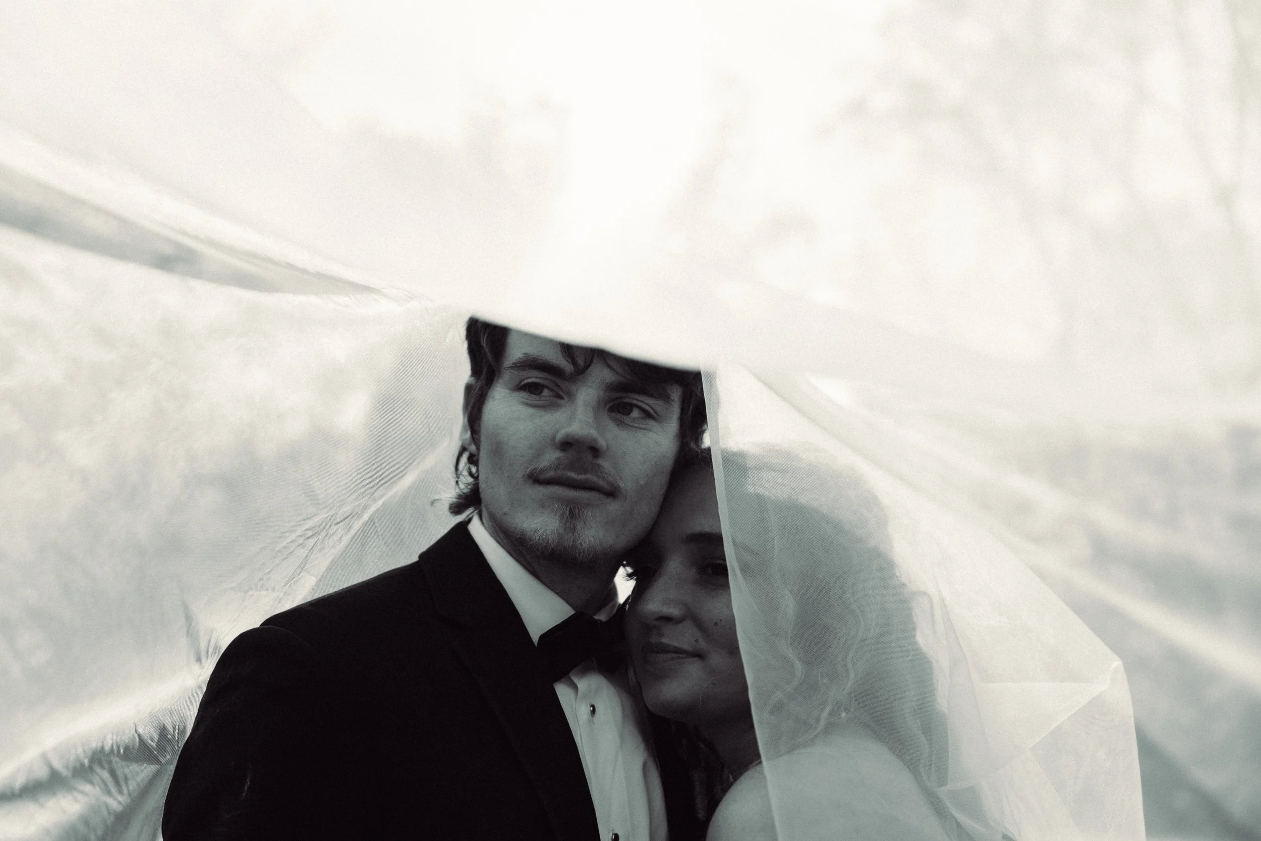 A black and white photo of a couple in wedding attire, standing under a veil. St. Louis Documentary-style wedding photography on 35mm film, 120 film, and digital by Trevor Lovatto